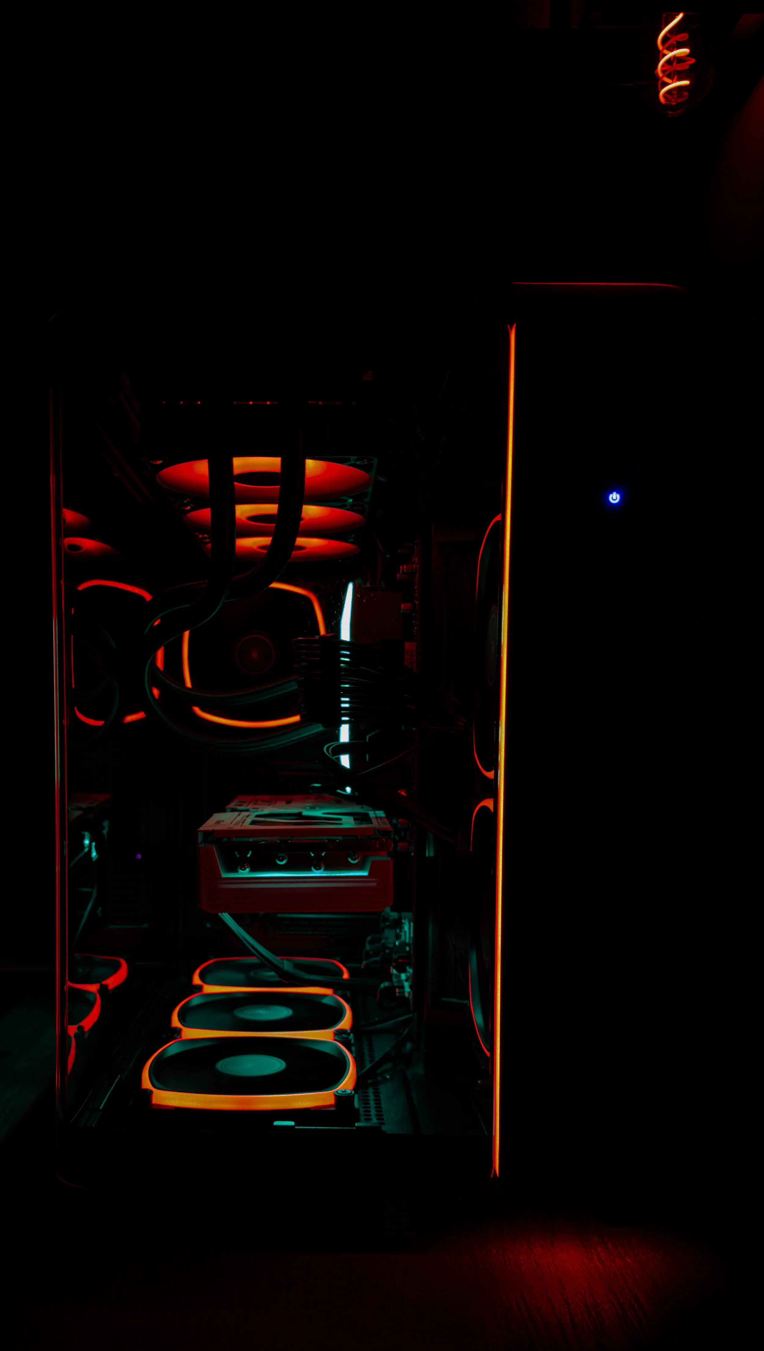 A darkened computer case with LED lighting showcasing internal components including cooling fans, a liquid cooling system with tubes, and other hardware components illuminated with red and blue lights.