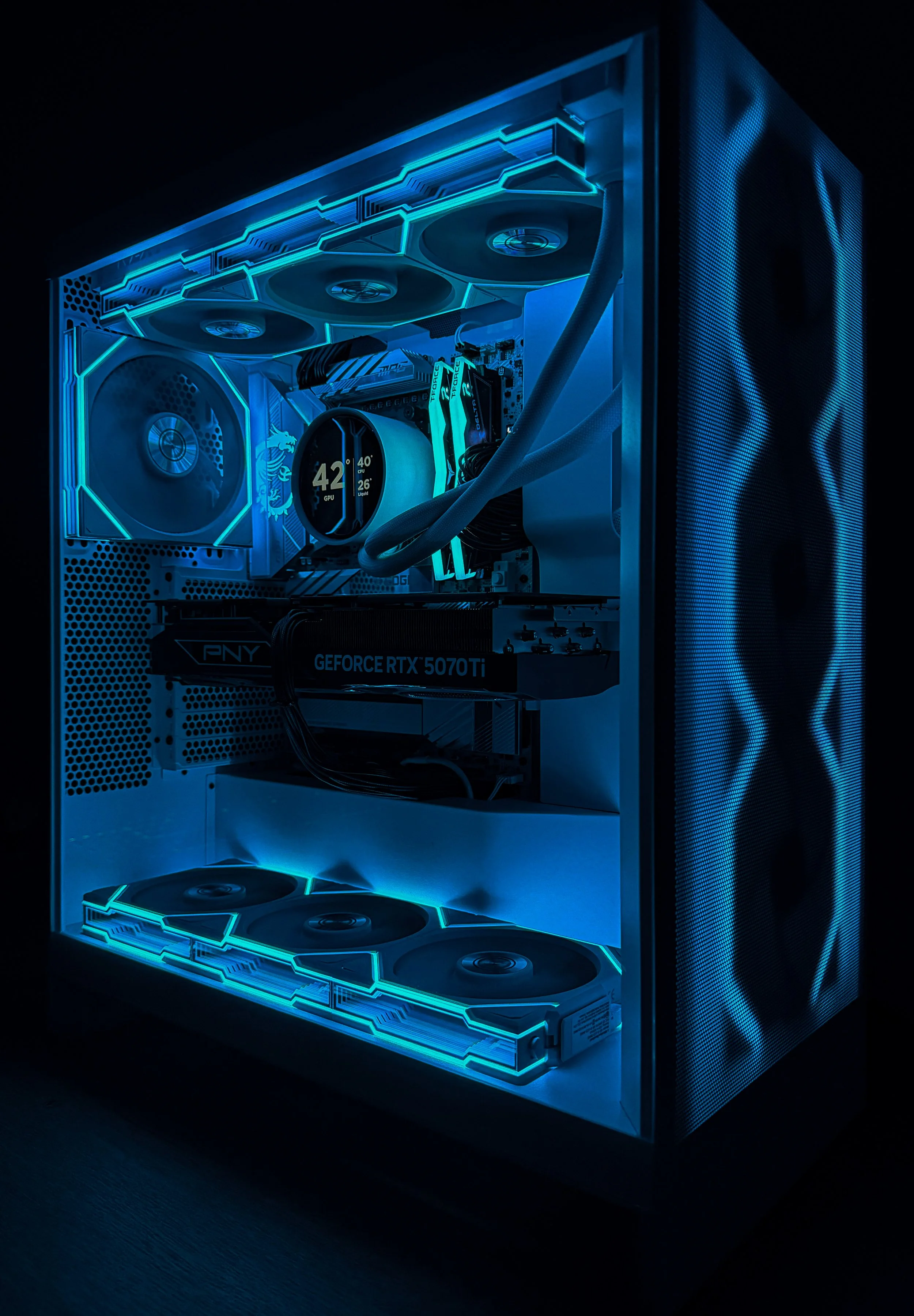 A gaming computer tower with illuminated interior components, including cooling fans, a graphics card labeled 'GEFORCE RTX 5070 Ti,' and a circular LED display showing temperature and GPU load in bright blue lighting.