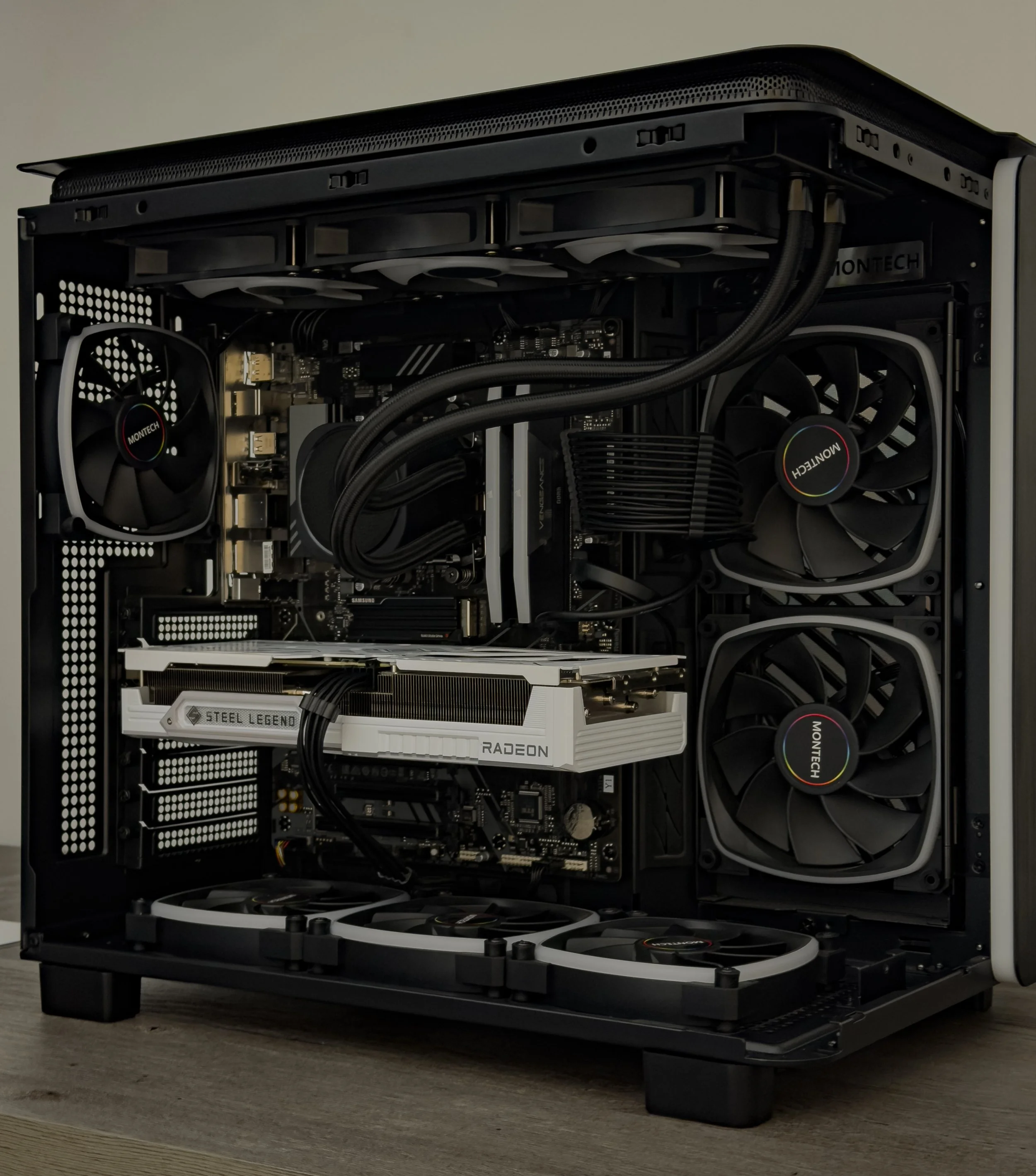 Open black computer case showing components: two large fans on top, a graphics card labeled 'Steel Legend Radeon', and several other internal hardware parts, all sitting on a wooden surface.