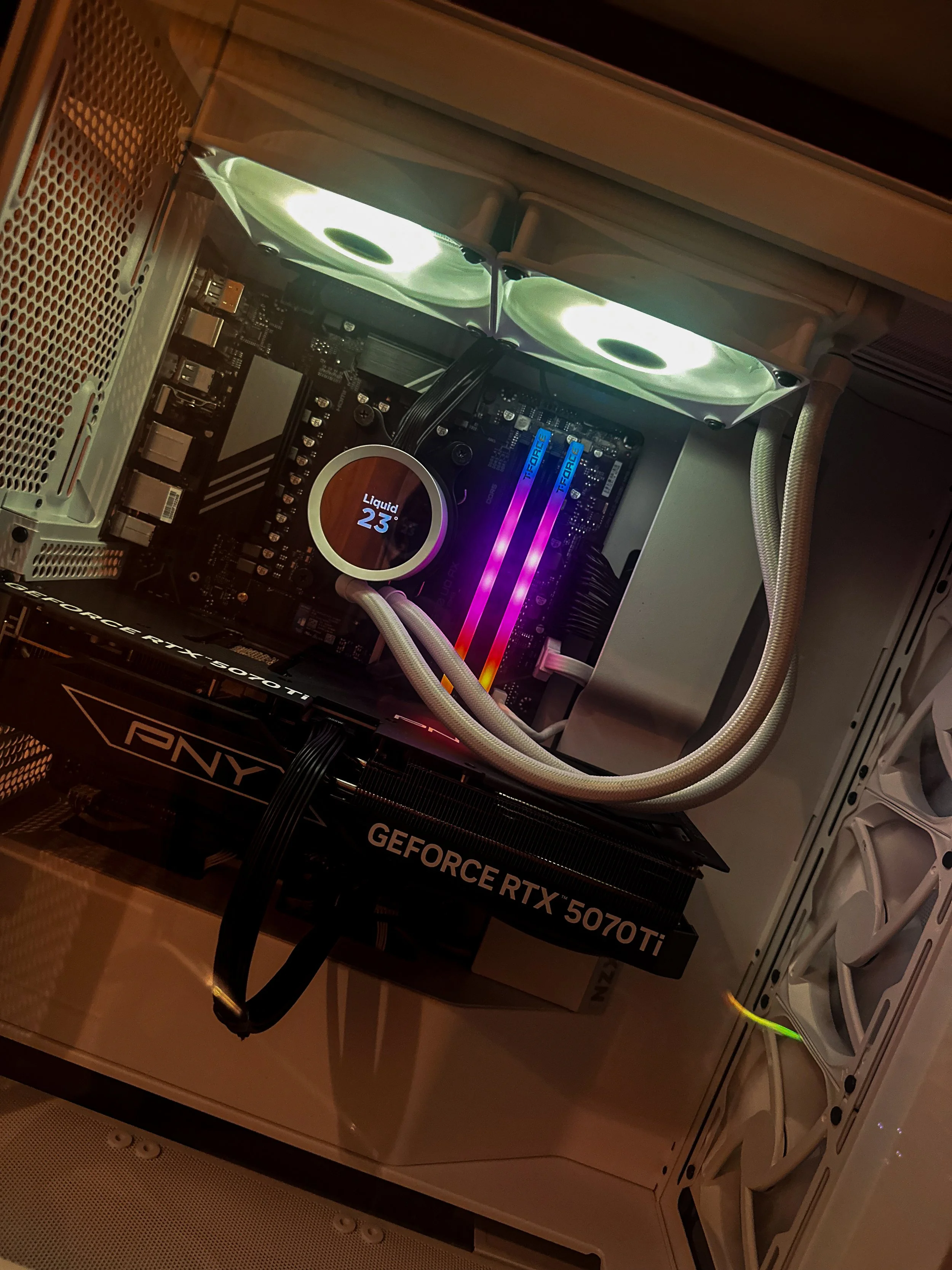Inside a computer case showing a graphics card, two cooling fans, RAM modules with RGB lighting, a motherboard with a liquid cooling system displaying 23 degrees Celsius, and various power and data cables.