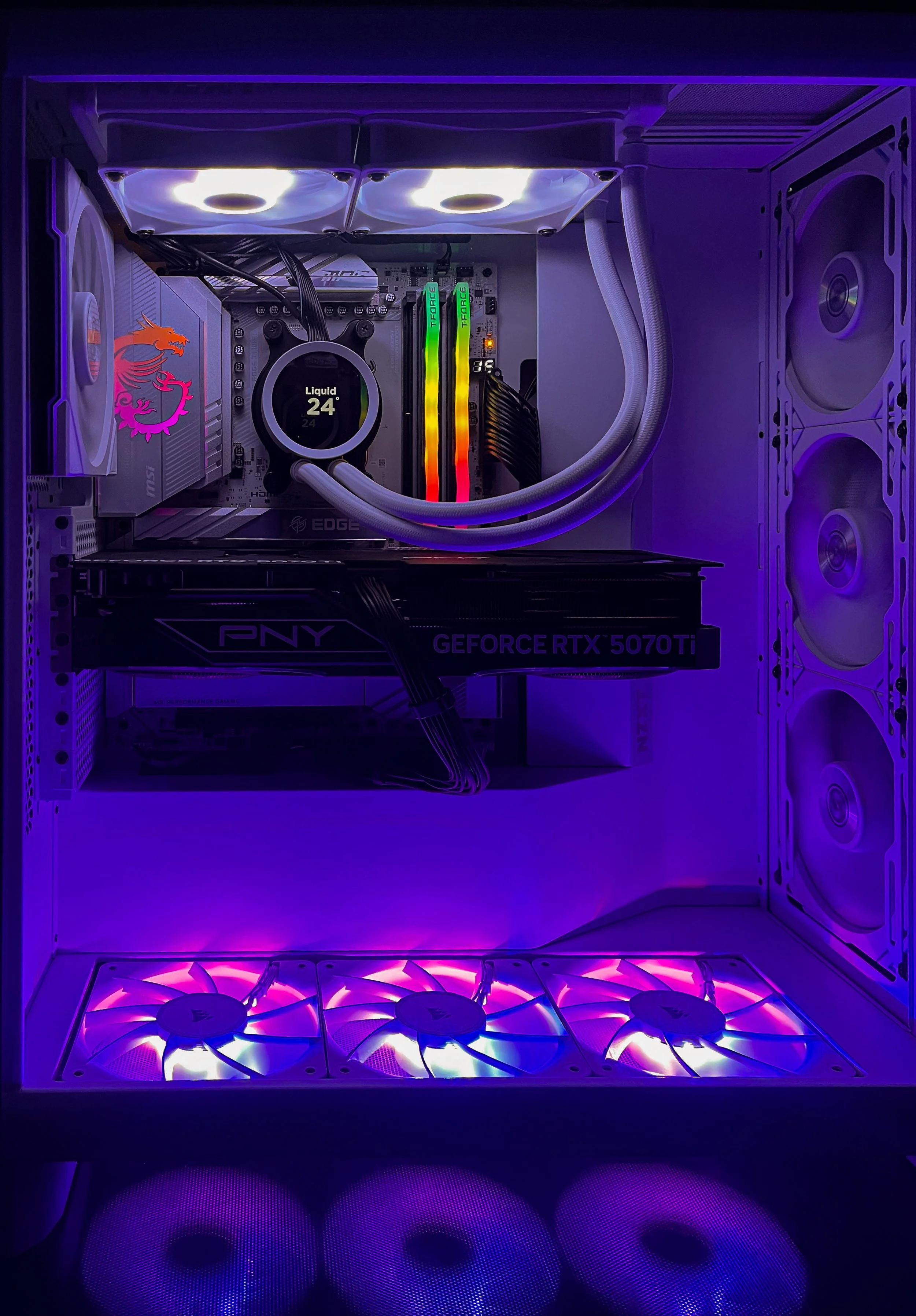 Inside a custom gaming PC with RGB lighting, showing cooling fans, a liquid cooling system with a digital temperature display, brightly colored RAM sticks, a graphics card labeled 'PNY GeForce RTX 5070 Ti', and illuminated white case components.