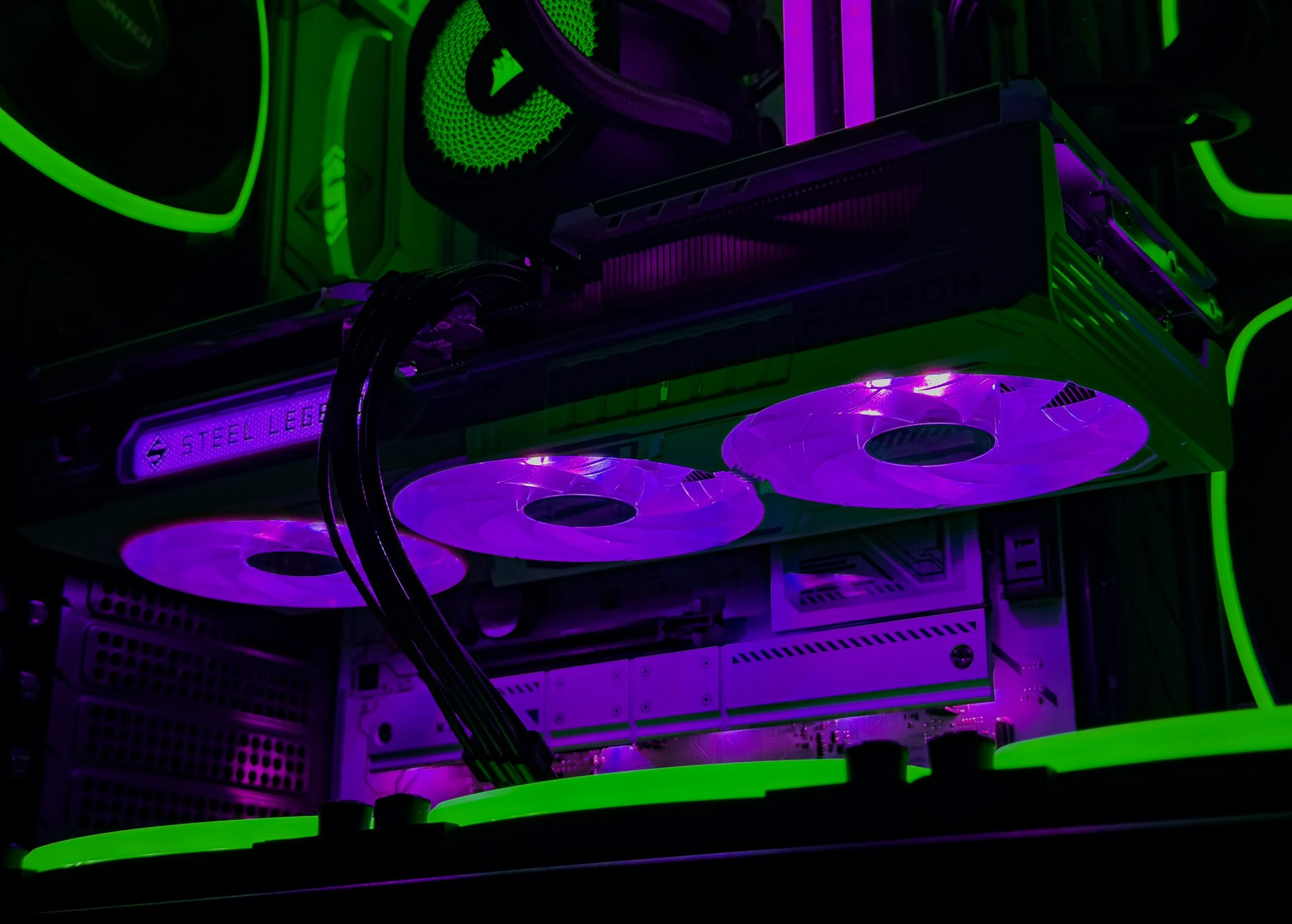 A close-up of a gaming computer's internal components illuminated with purple and green LED lights, showing a graphics card, cooling fans, and various cables.