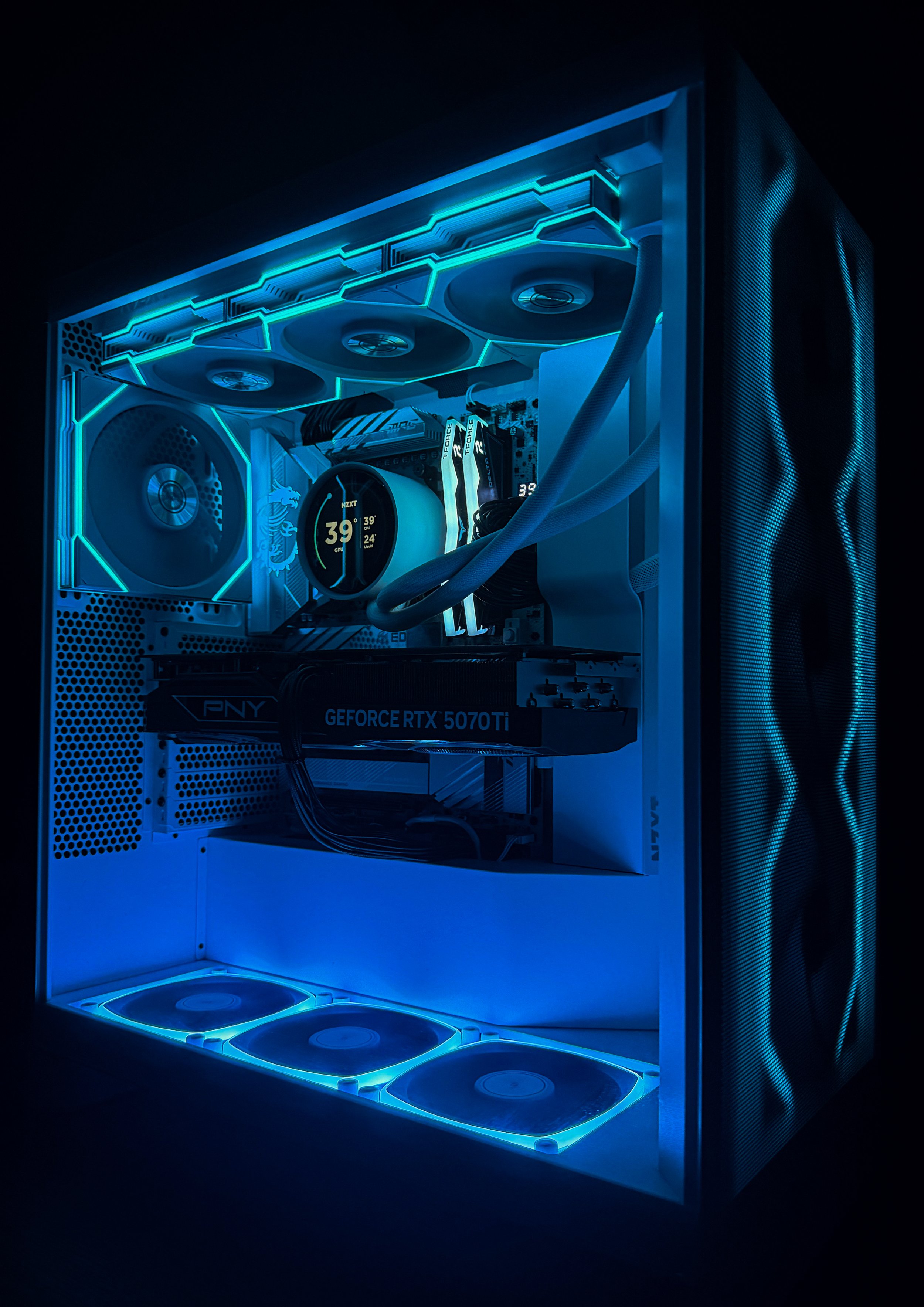 A custom gaming computer with white case interior illuminated by blue LED lighting, featuring three cooling fans, a liquid cooling system, RGB RAM sticks, a large graphics card labeled 'GEFORCE RTX 5070Ti,' and a temperature monitor display showing 3