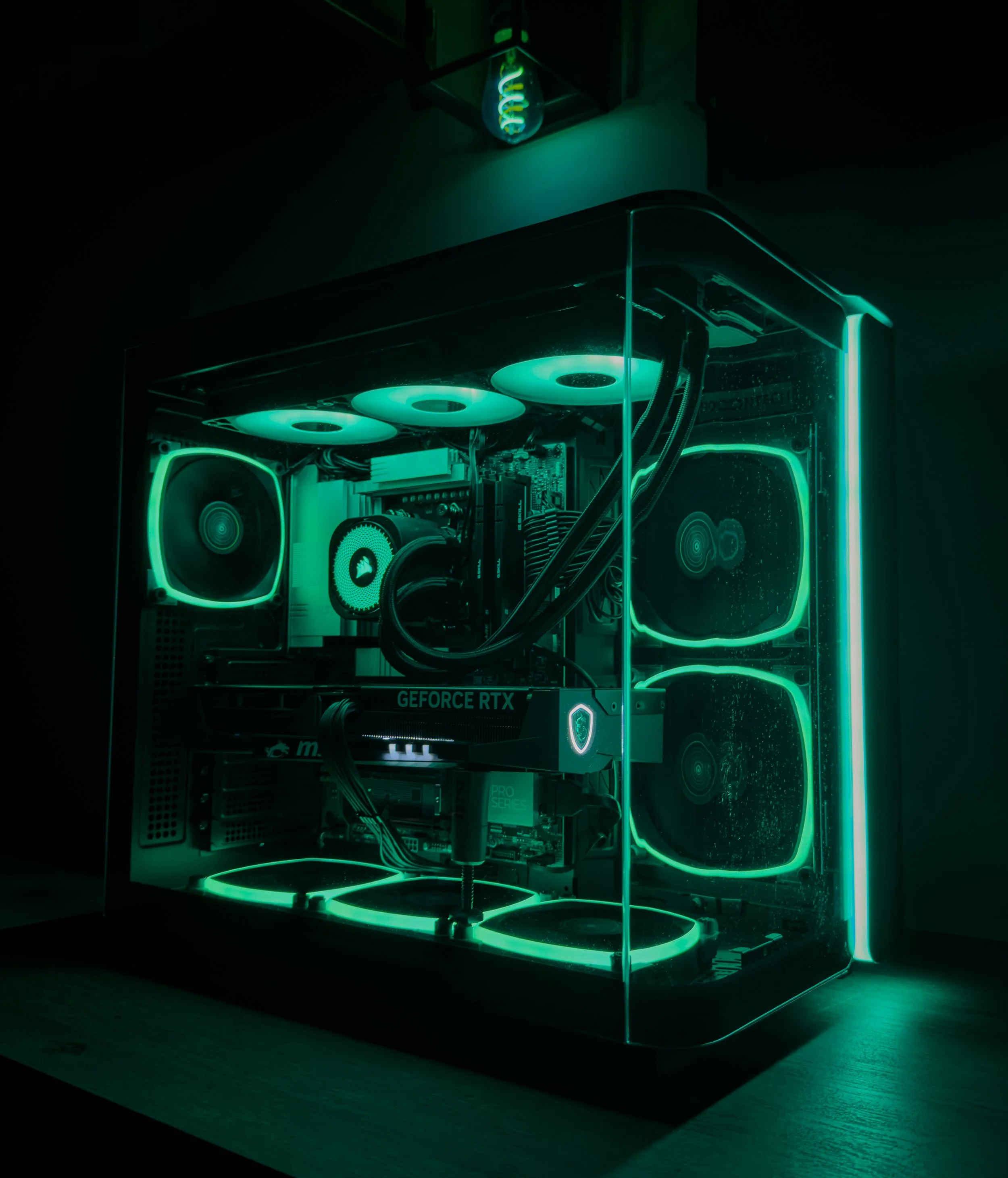 A gaming computer with a transparent case, illuminated by green LED lights, placed on a wooden desk with a glass side panel and internal cooling fans visible.
