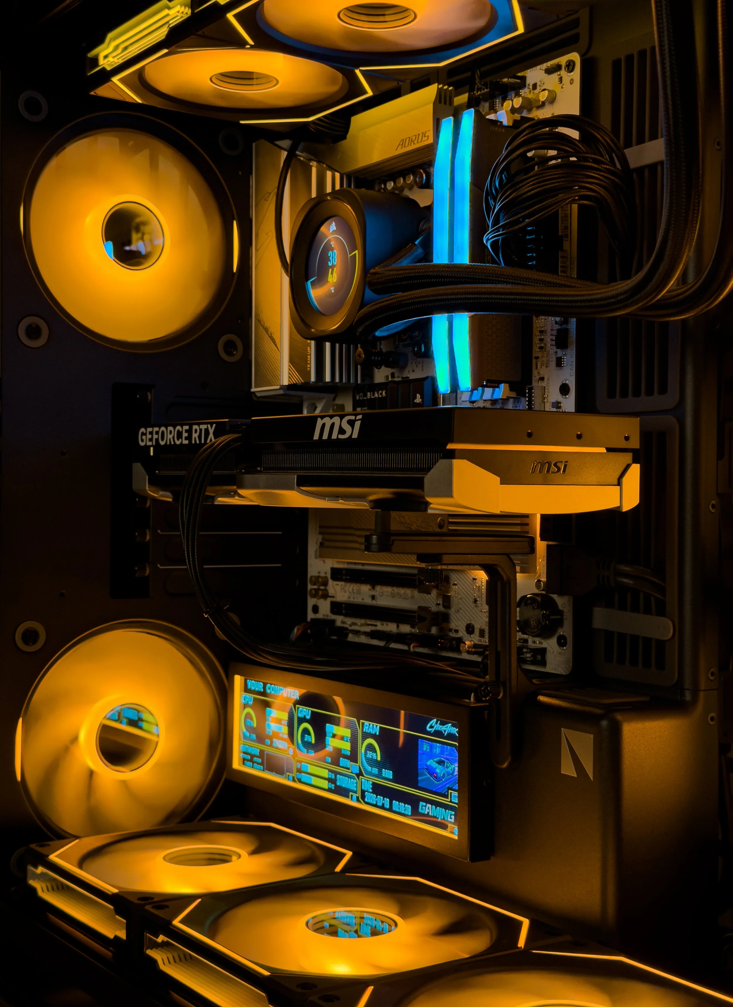 Inside a custom gaming computer with RGB lighting, a large circular fan on the left, an MSI graphics card, a motherboard, RAM sticks, a digital display showing computer stats, and yellow LED accents.