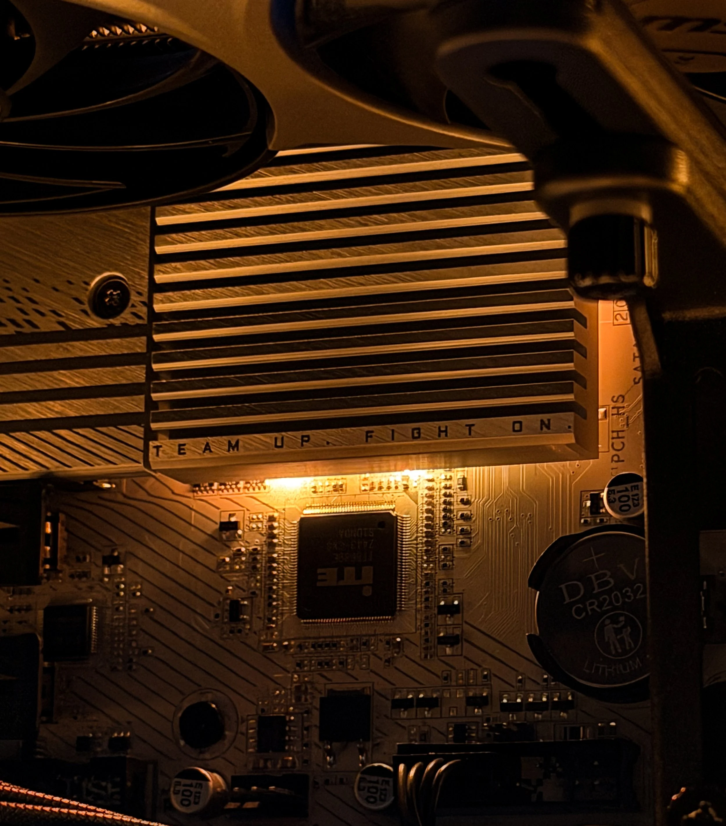 Close-up of a computer motherboard with various electronic components including a circuit board, capacitors, and wiring, illuminated by warm light.