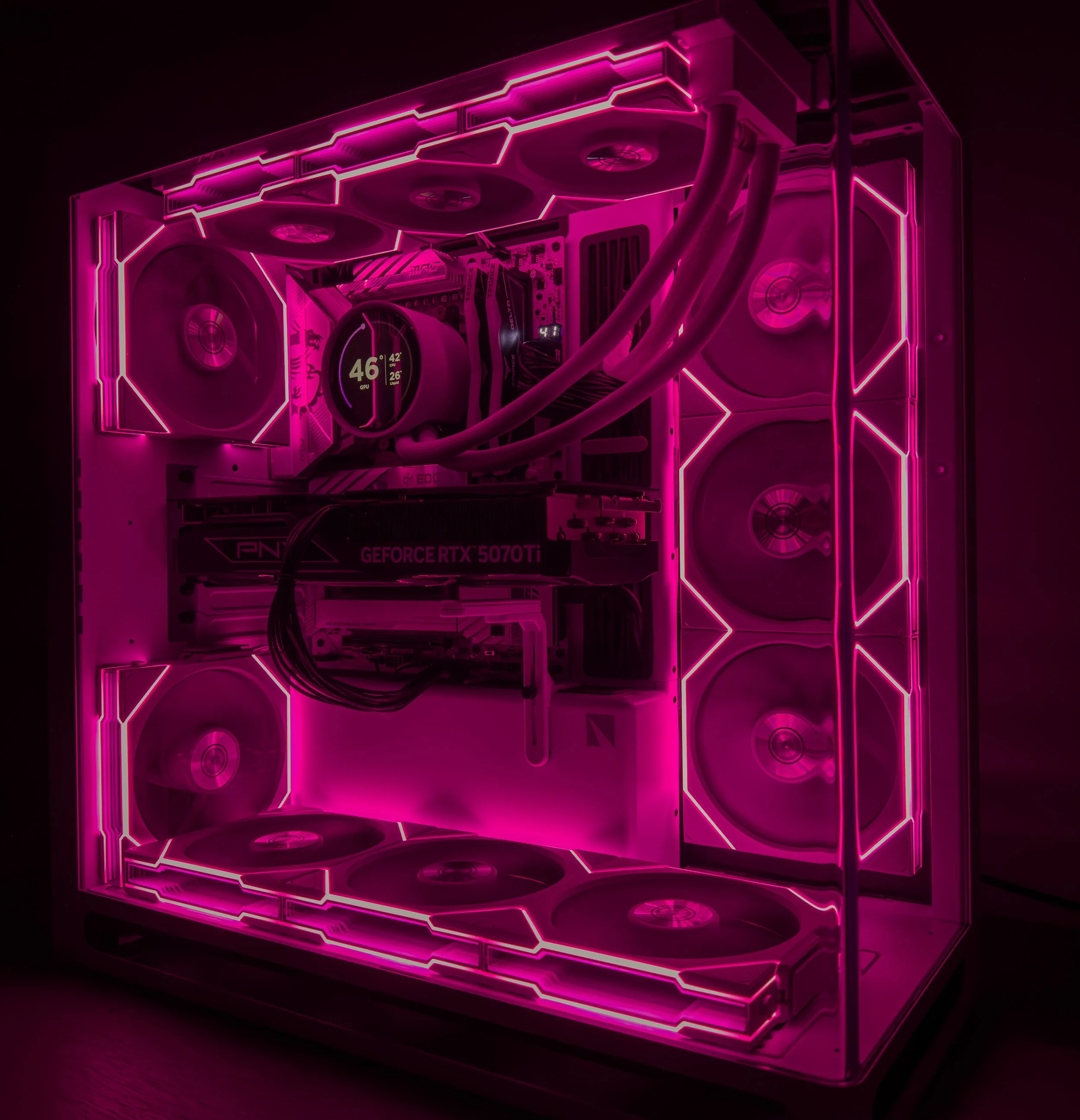 A close-up view of a custom-built gaming computer with a transparent side panel illuminated by pink neon lighting. The interior features a high-performance graphics card, cooling fans, and a digital temperature display.
