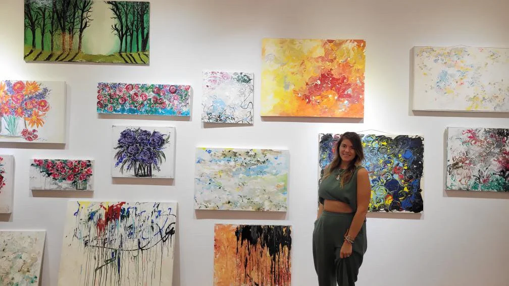 A woman stands in front of a white wall with various colorful abstract paintings and floral artworks displayed.