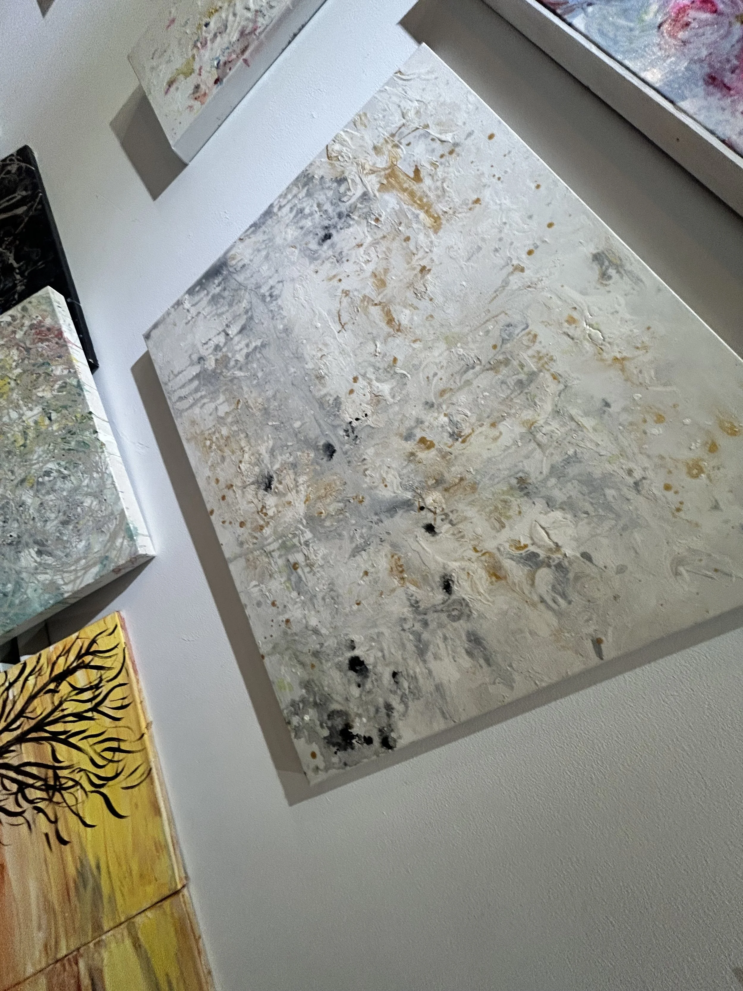 Several abstract paintings are displayed on a wall. The paintings have various colors and textures, with one featuring a tree-like black design on a yellow background, and others with splashes of color and textured surfaces.