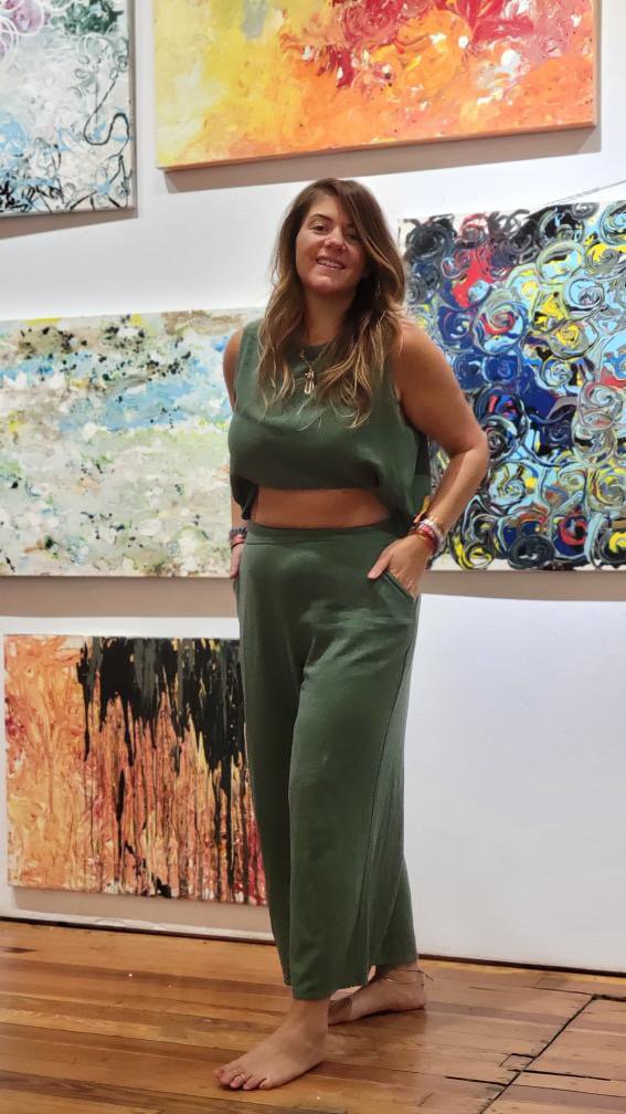 A young woman with long brown hair and a smile, standing barefoot in an art gallery, wearing a green sleeveless top and matching loose pants. She has bracelets on her left wrist and is posing in front of colorful abstract paintings hanging on the wall.