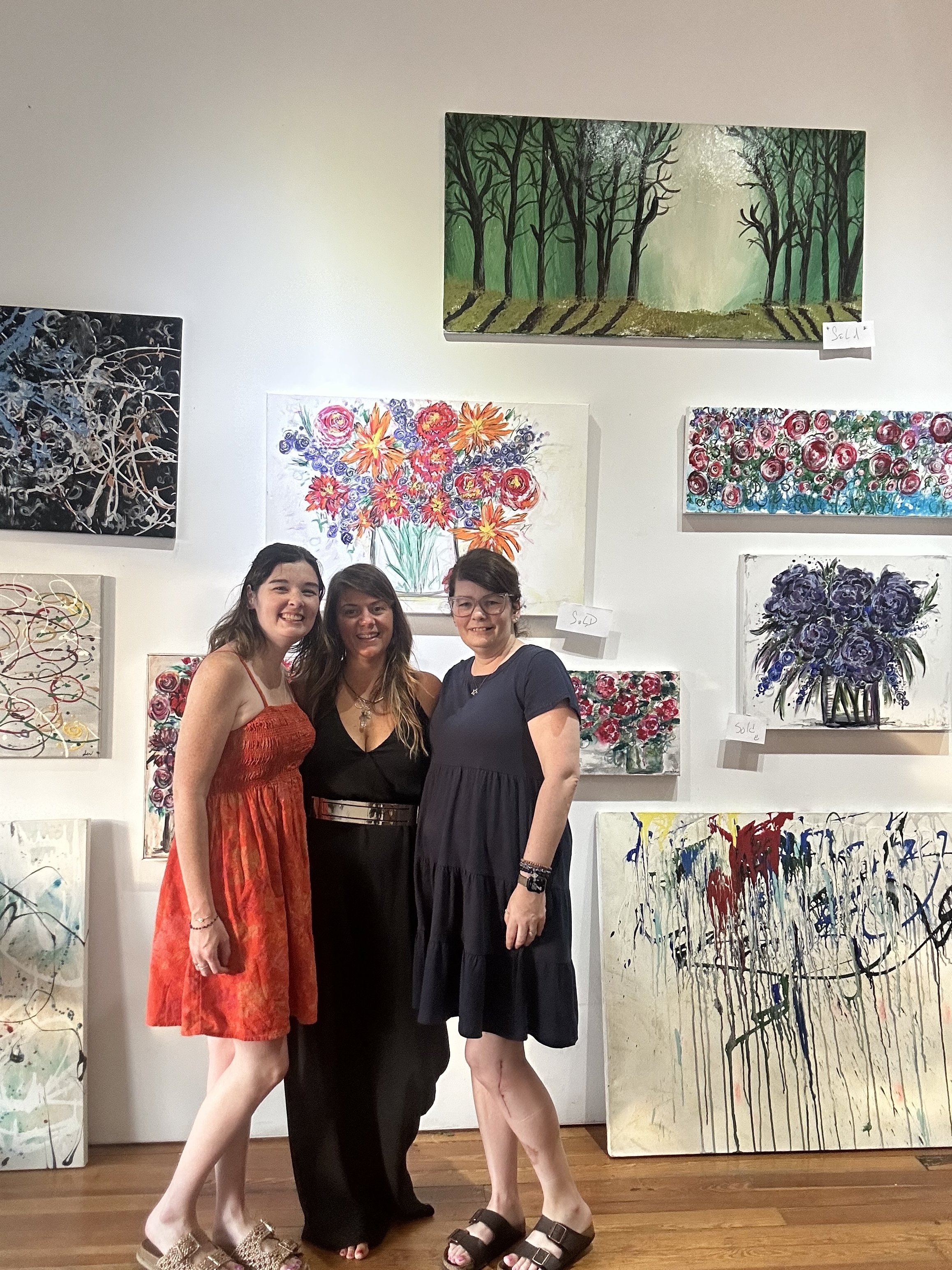Three women standing in an art gallery in front of colorful abstract paintings, smiling at the camera.