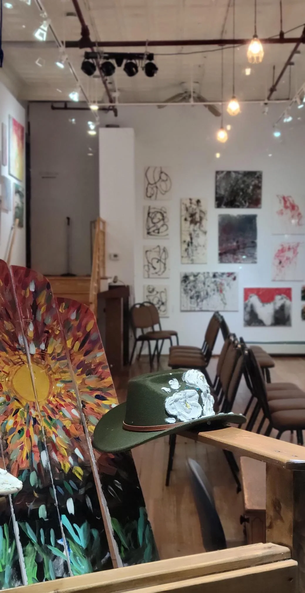 An art gallery with paintings on the wall, chairs arranged, and a green hat with white flowers on a wooden bench in the foreground.