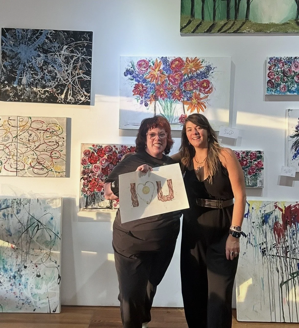 Two women smiling and standing in front of a wall of colorful abstract and floral paintings at an art gallery. One woman holds a sign with a heart-shaped fried egg and bacon that spells 'I U'.