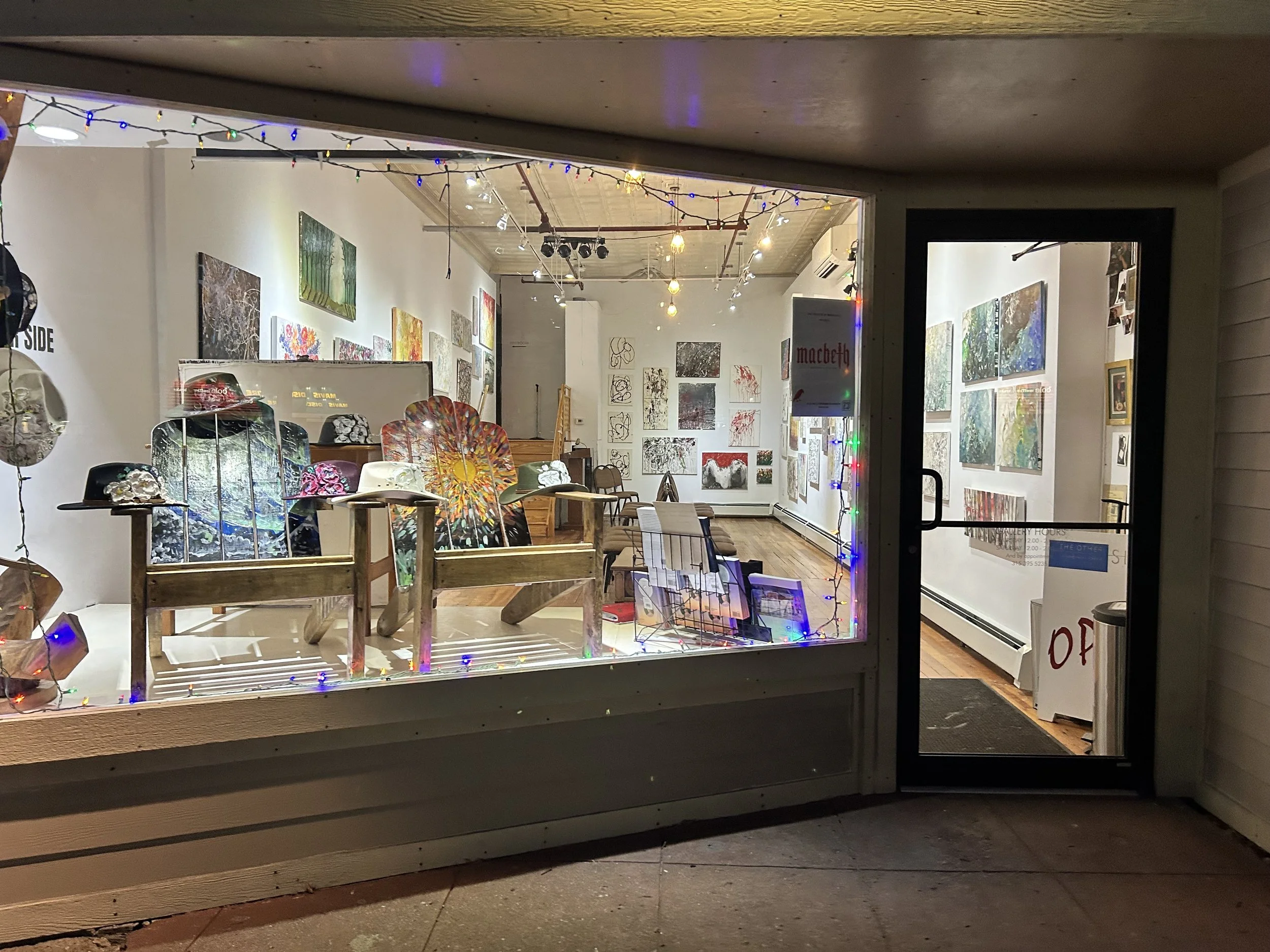 View of an art gallery interior through a storefront window. The gallery features colorful paintings on the walls, with some displayed on easels and tables in the foreground. The gallery is decorated with string lights and has a wooden floor.
