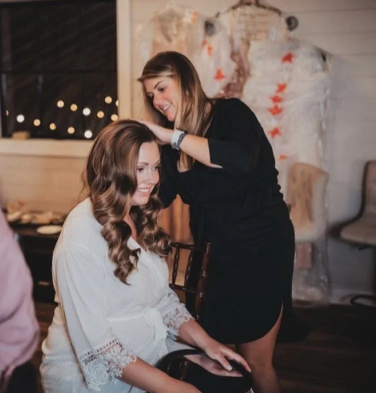Two women, one seated with long, curly brown hair and a white lace-topped robe, and the other standing with straight, shoulder-length brown hair, smiling and touching the seated woman's head, in a warmly lit room with wooden decor and fairy lights.