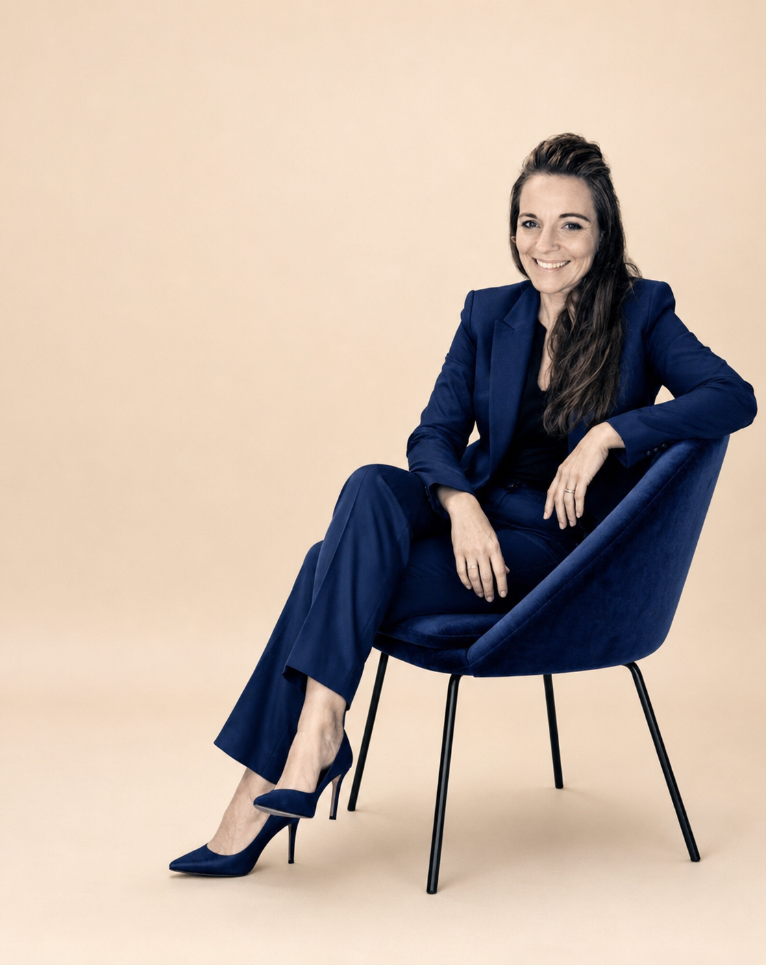 A woman with long brown hair dressed in a blue suit, sitting on a blue chair, smiling at the camera against a beige background.