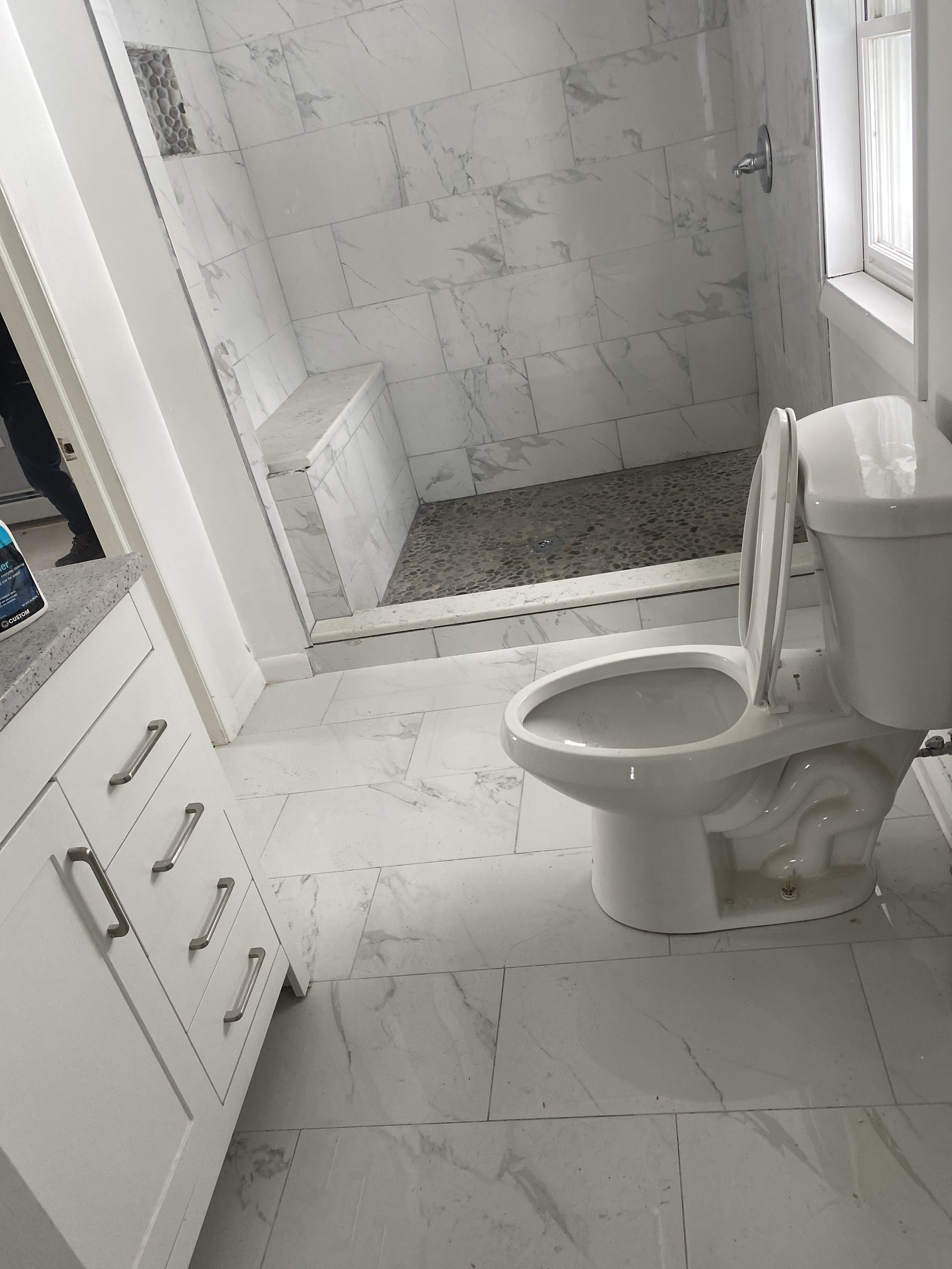 A bathroom with white marble tiles on the walls and floor, a white toilet with its lid open, a marble bench inside the shower, and a window providing natural light.