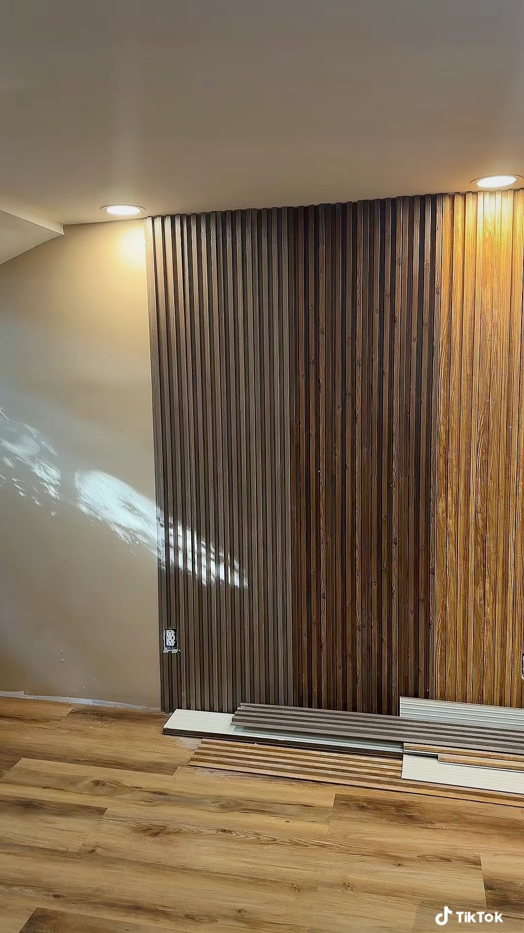 Interior of a room with a partially installed wooden wall panel, some planks on the floor, and two ceiling lights.