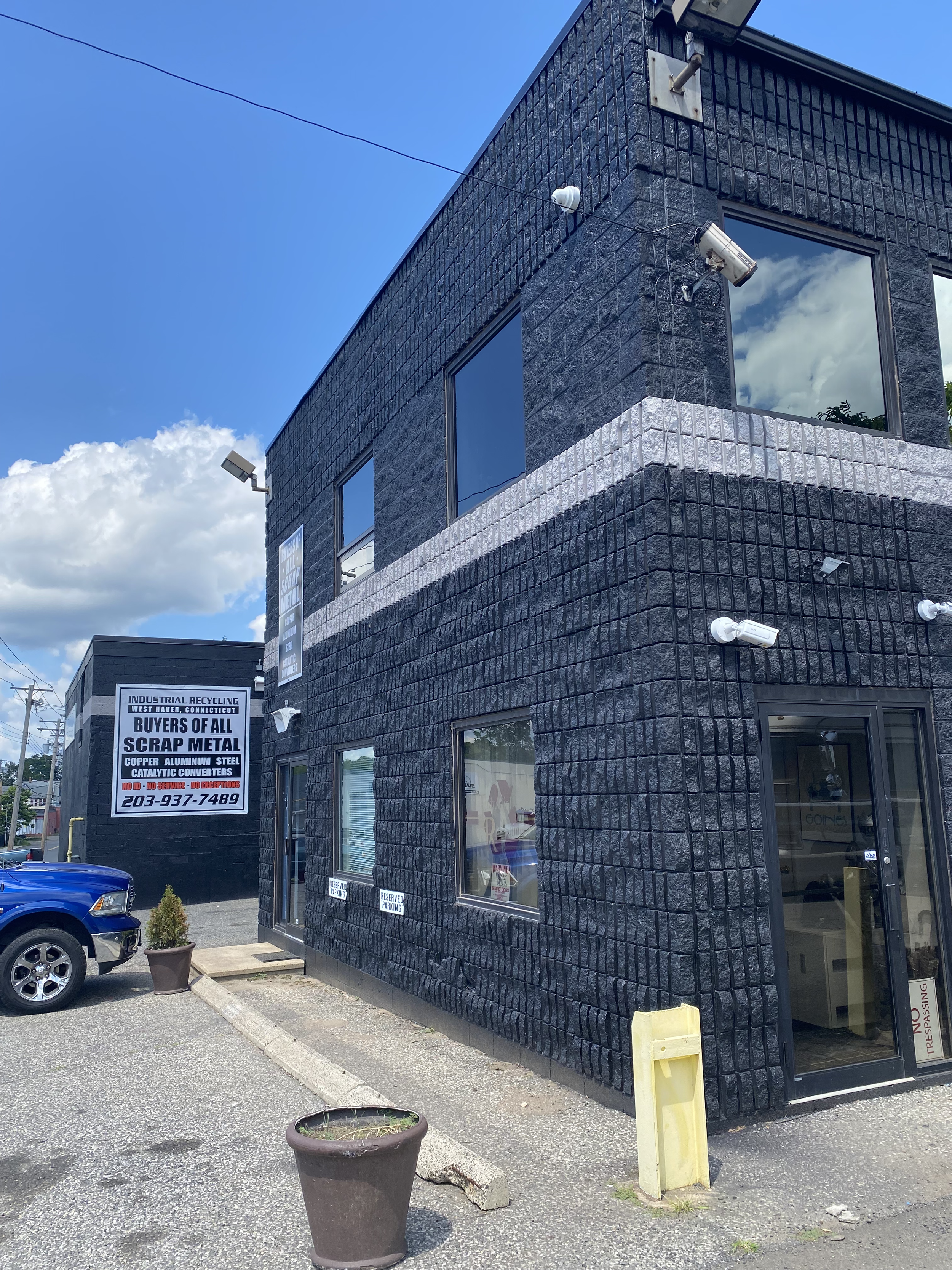 The exterior of a black brick commercial building with large glass windows, security cameras, and a signboard for a scrap metal recycling business, adjacent to a parking lot with a blue vehicle and a large potted plant.