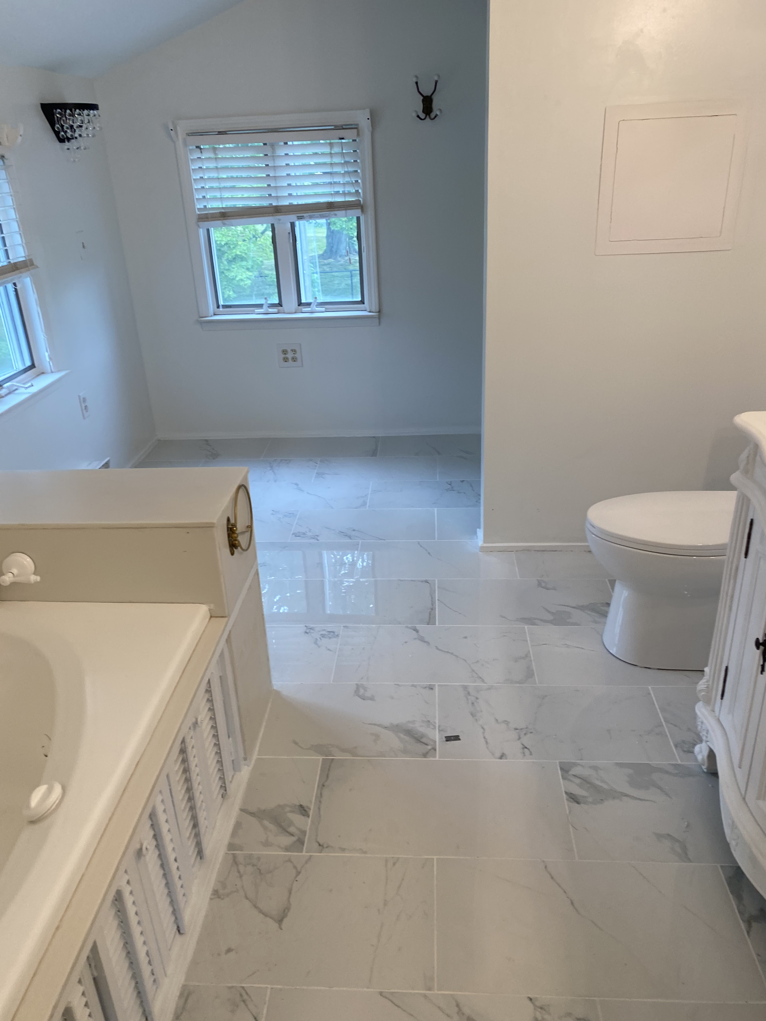 A bathroom with white walls and marble floor tiles. There is a beige vanity with louvered doors on the left side and a toilet on the right. Two windows with white blinds are visible, and there are wall-mounted light fixtures and a decorative wall hoo