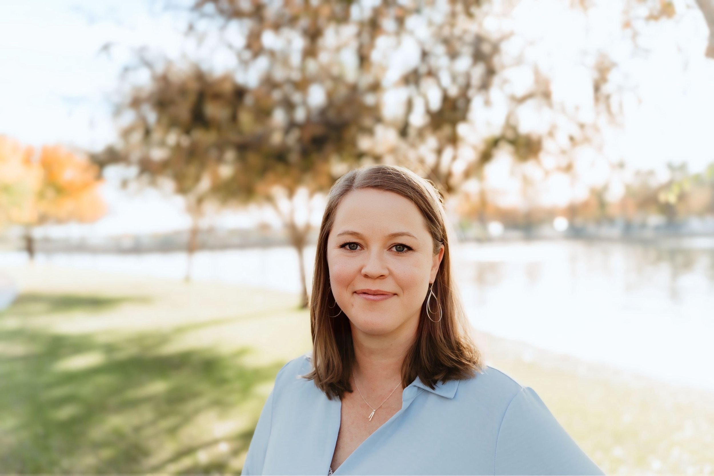 A young woman with shoulder-length hair and earrings, wearing a light blue button-up shirt, standing outdoors with trees and a body of water in the background. It is a sunny day with clear skies.