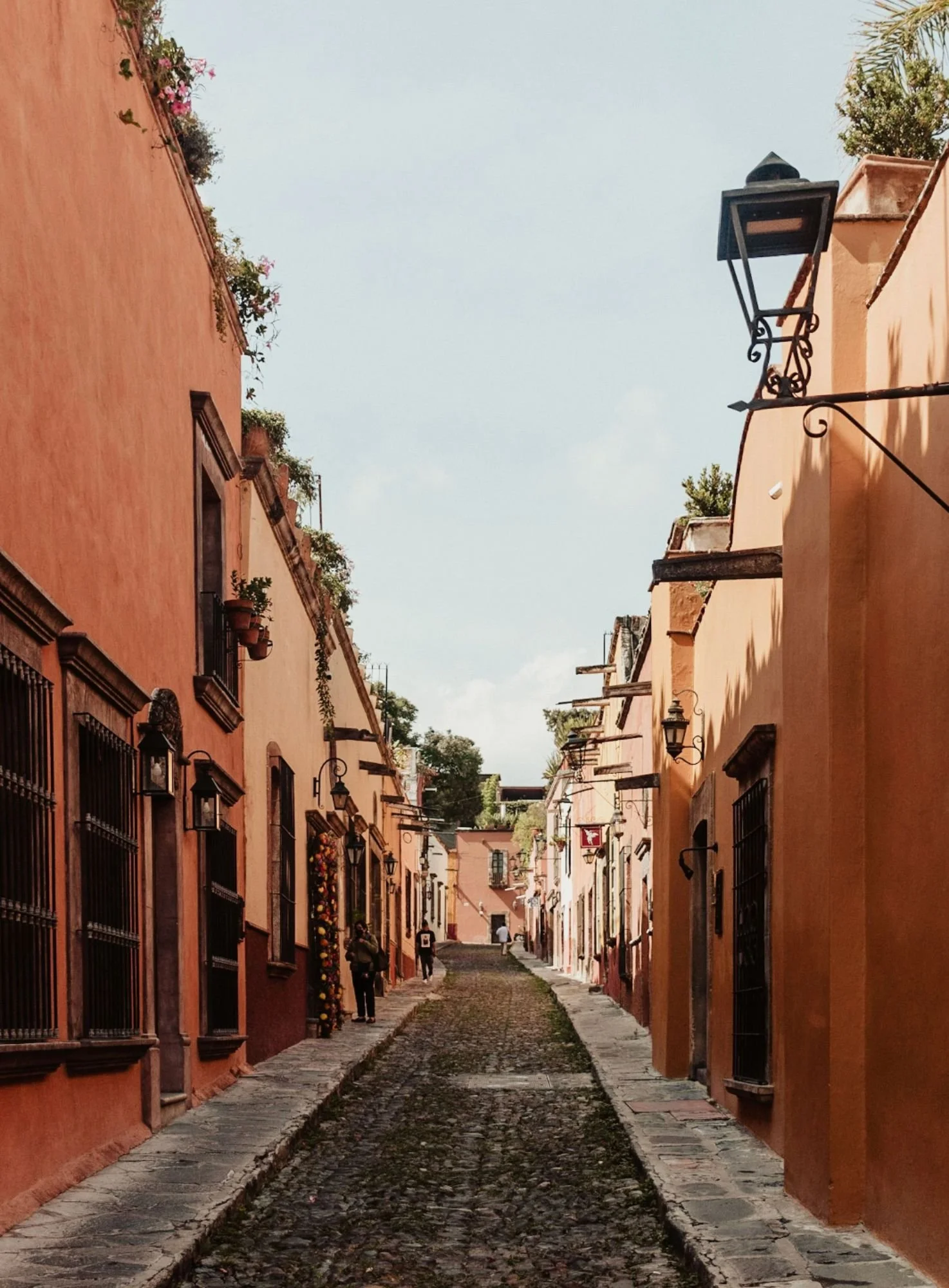 A narrow cobblestone street lined with pink and orange buildings, featuring black iron window bars, hanging lanterns, and potted plants, with a few people walking in the distance under a partly cloudy sky.