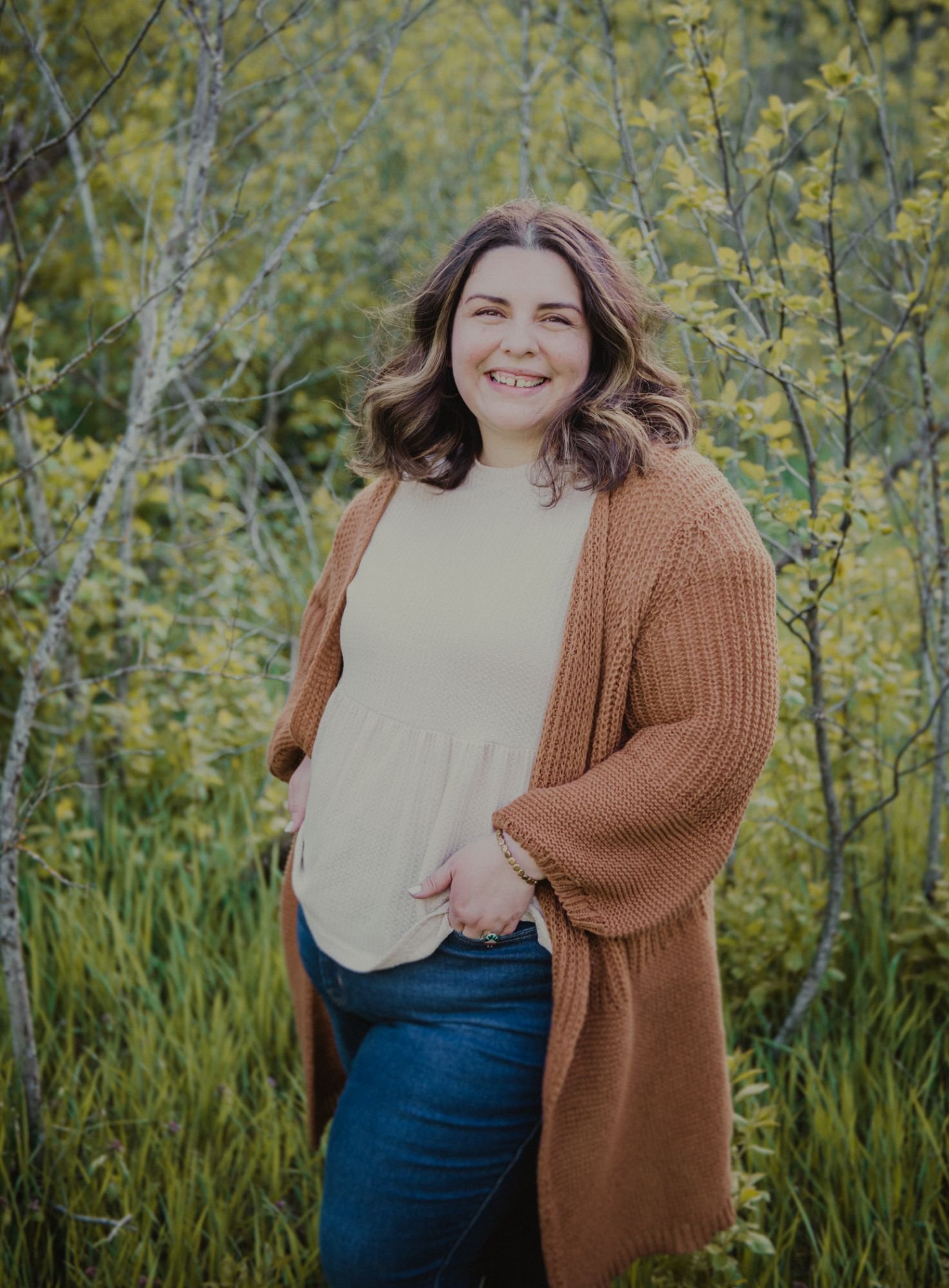 A woman smiling outdoors in a park with green and yellow trees and grass in the background. She is wearing a beige top and a brown cardigan, with blue jeans, and has shoulder-length wavy brown hair.
