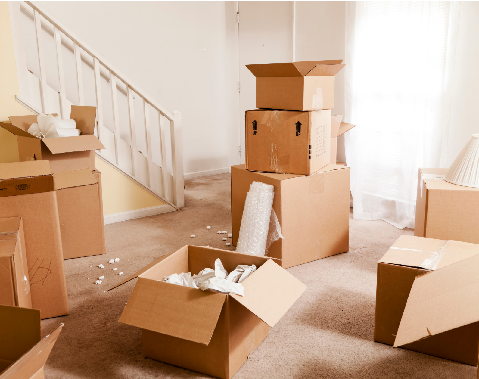 A cluttered living room with several open cardboard moving boxes, some filled with packing materials and paper, on a brown carpet. A staircase with white railing is in the background, and sunlight filters through sheer white curtains.