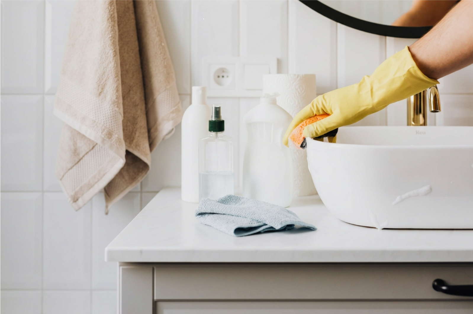 Person cleaning a white sink with a yellow glove, using a sponge, on bathroom countertop with soap bottles and towels.