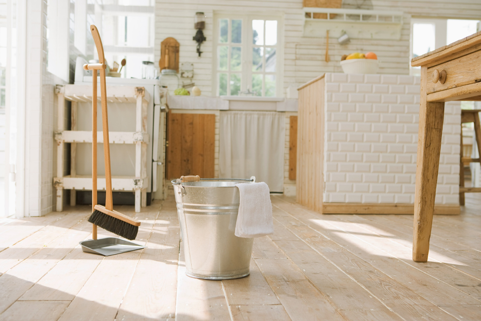 Cleaning tools including a broom, dustpan, and bucket with a towel in a bright, rustic kitchen with wooden flooring and white brick and wood accents.