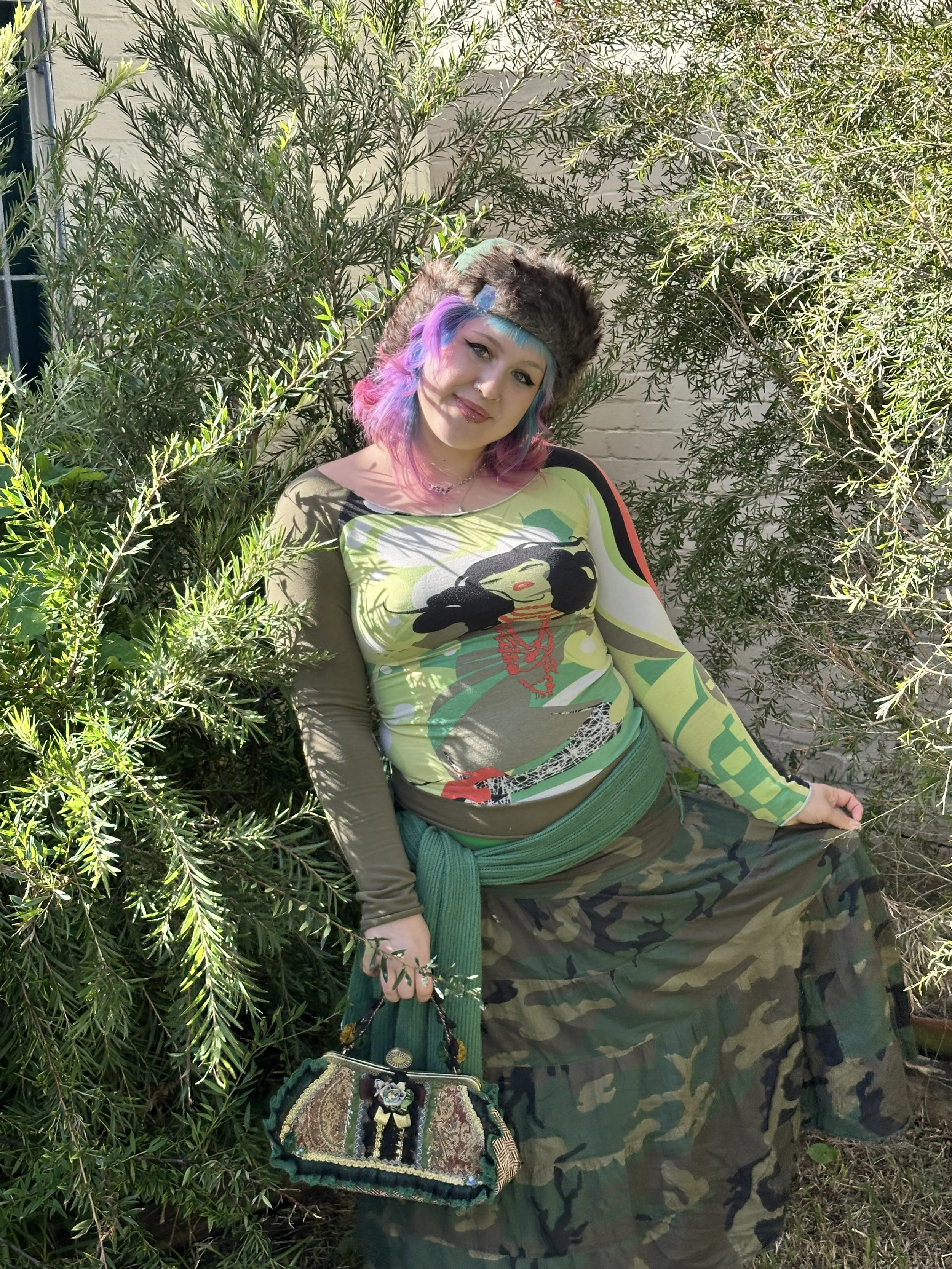 A woman with multicolored hair sitting outdoors among green bushes. She is wearing a fur hat, a long sleeve shirt with a graphic print, camouflage pants, a green belt, and is holding a small decorated bag.