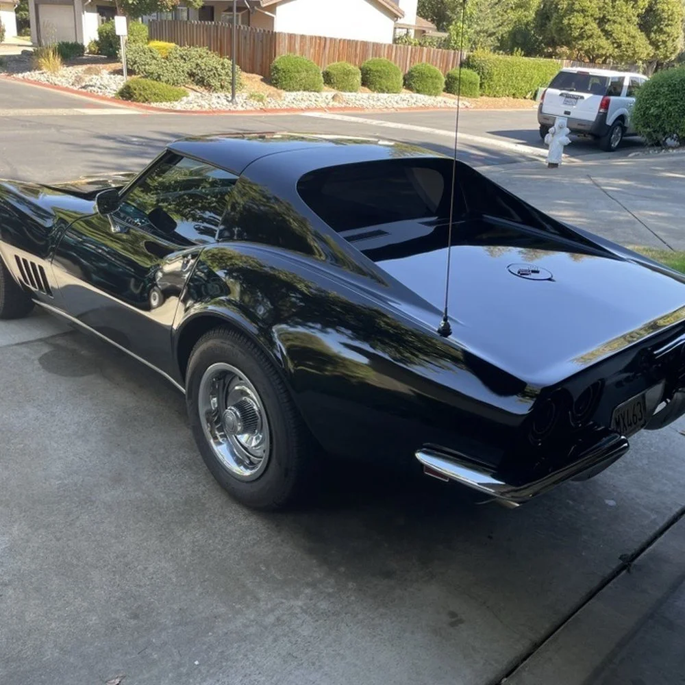 A black vintage Corvette sports car parked on a driveway in a suburban neighborhood, with houses, trees, and other parked cars visible in the background.