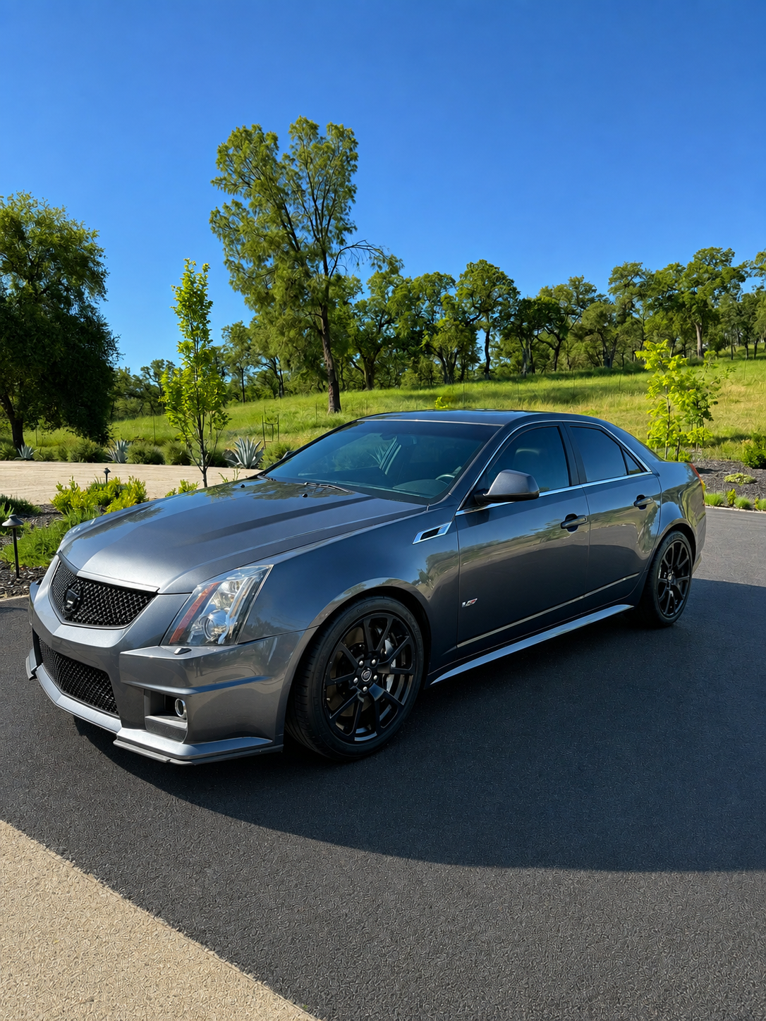 Gray Cadillac sedan parked on a paved driveway with green trees and grass in the background under a clear blue sky.