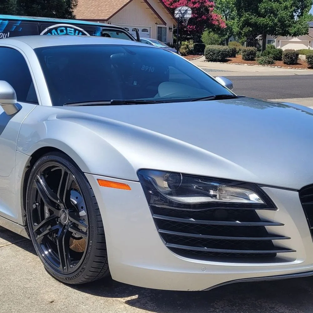 A silver Audi sports car parked in a suburban driveway with black wheels and tinted windows, with other vehicles visible in the background.