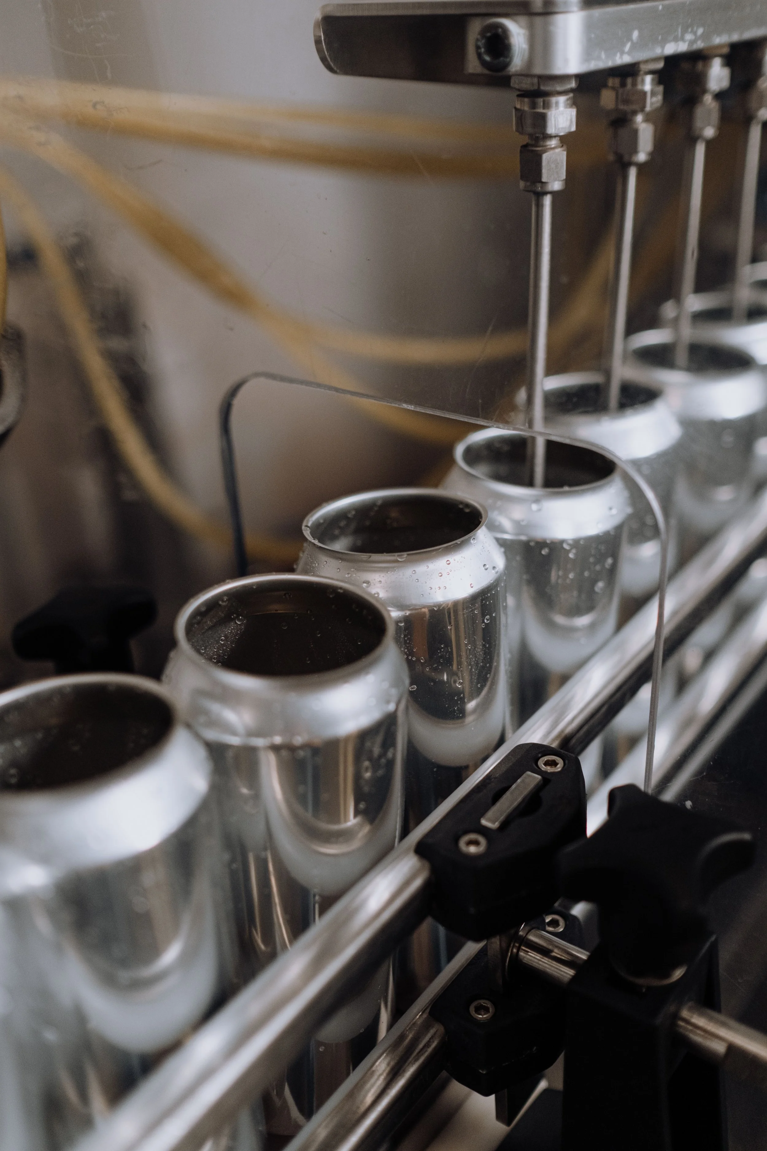 A metal machine filling aluminum cans with liquid, likely part of a manufacturing or bottling process, with visible pipes, bolts, and a metal frame.