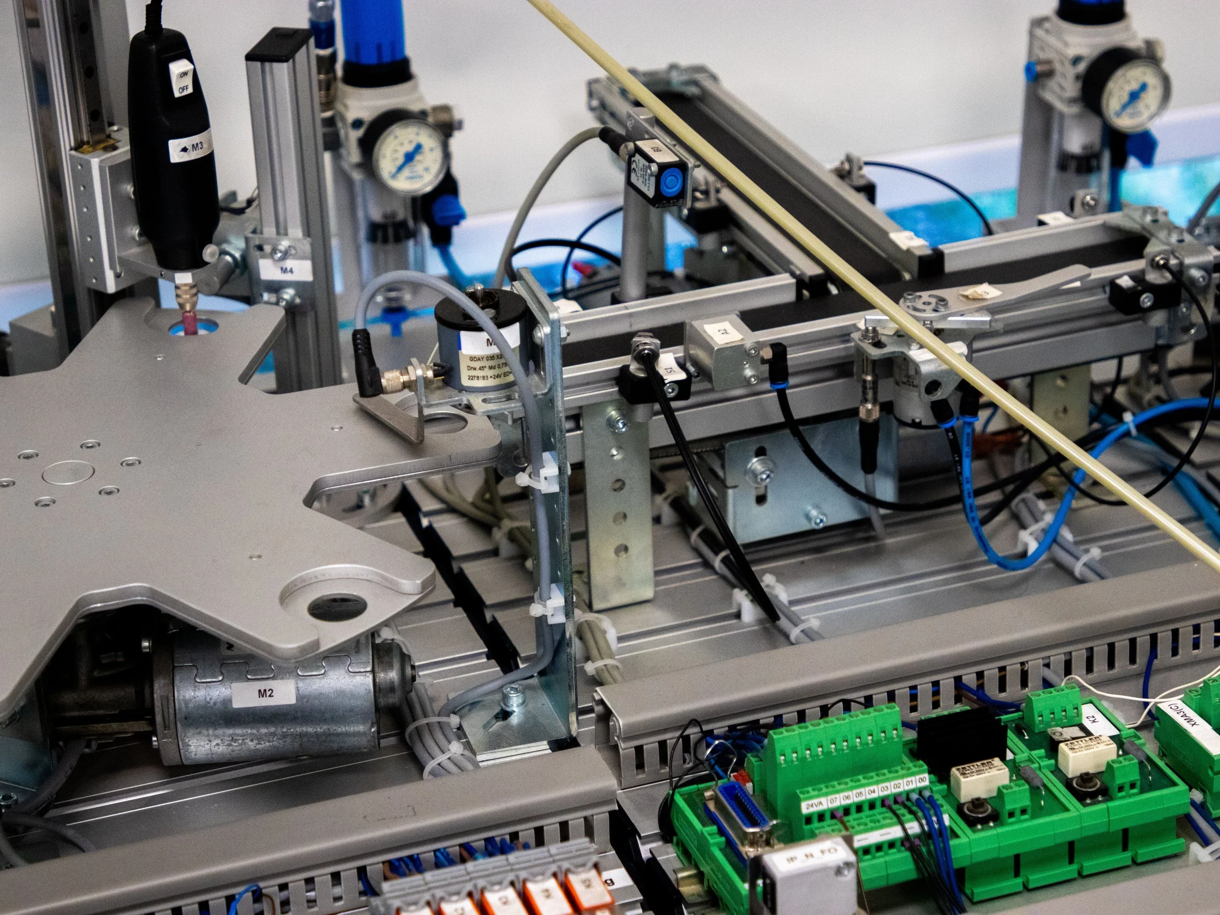 Close-up view of an industrial automation system with metal frame structure, pneumatic cylinders, valves, wiring, and green terminal blocks.