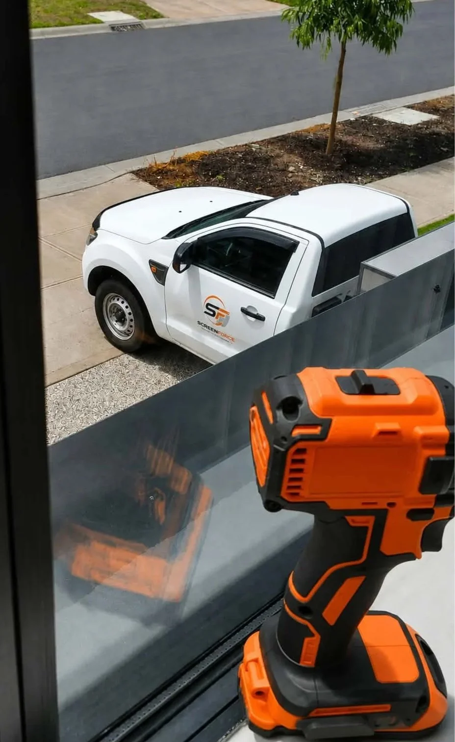 View from inside a house showing an orange cordless drill on a windowsill, a white pickup truck with a 'Screen Force' logo parked outside, a tree, and a paved street.