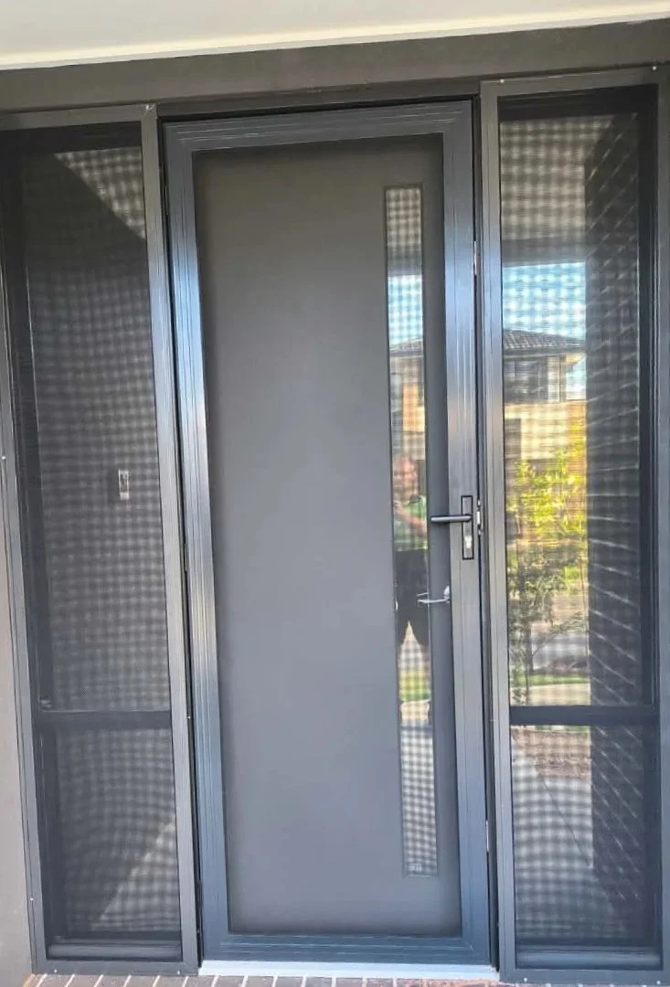 A glass door with a metal frame, flanked by sliding screens on both sides, leading to an outdoor area with a house, trees, and a lawn visible through the glass.