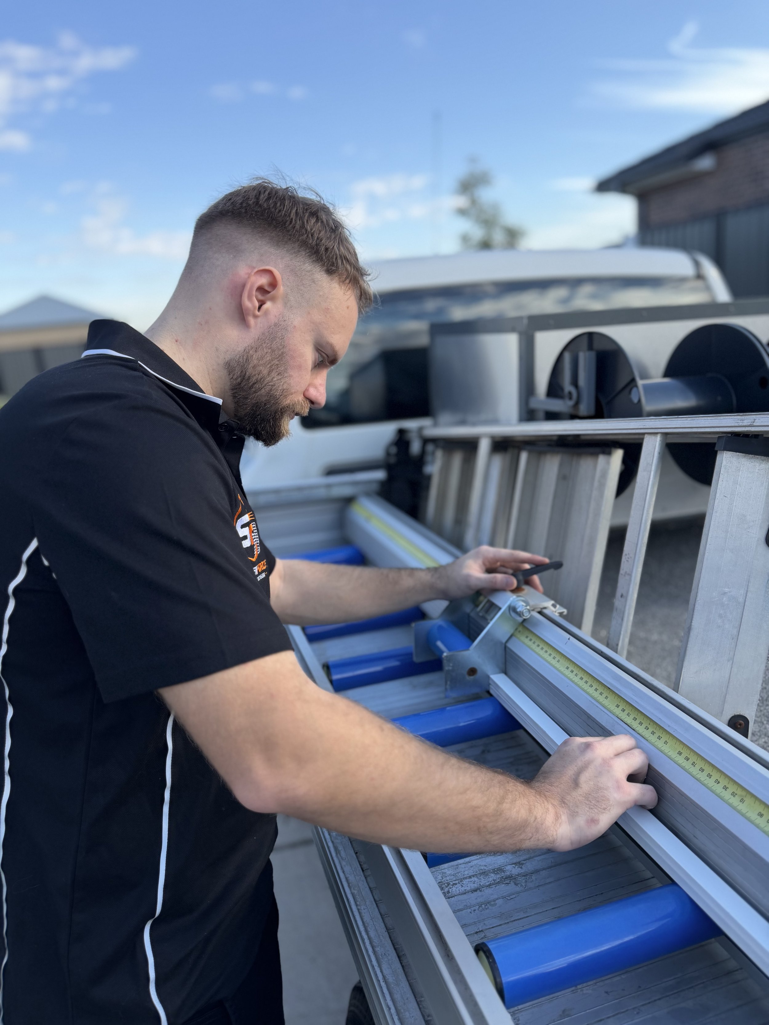 A man with a beard wearing a black shirt working on a piece of equipment outdoors during daytime with a blue sky and some clouds.