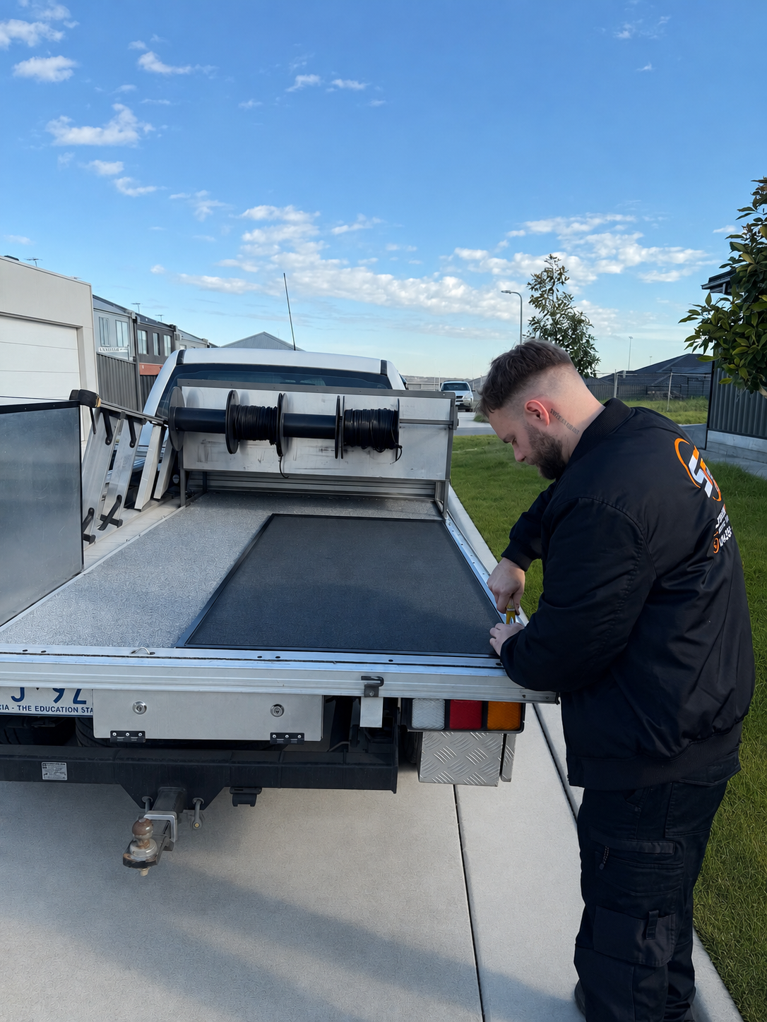 A man in a black jacket is working on the back of a utility truck with a flatbed, picking up tools or equipment. The truck is parked on a residential street with modern houses, greenery, and a partly cloudy blue sky in the background.