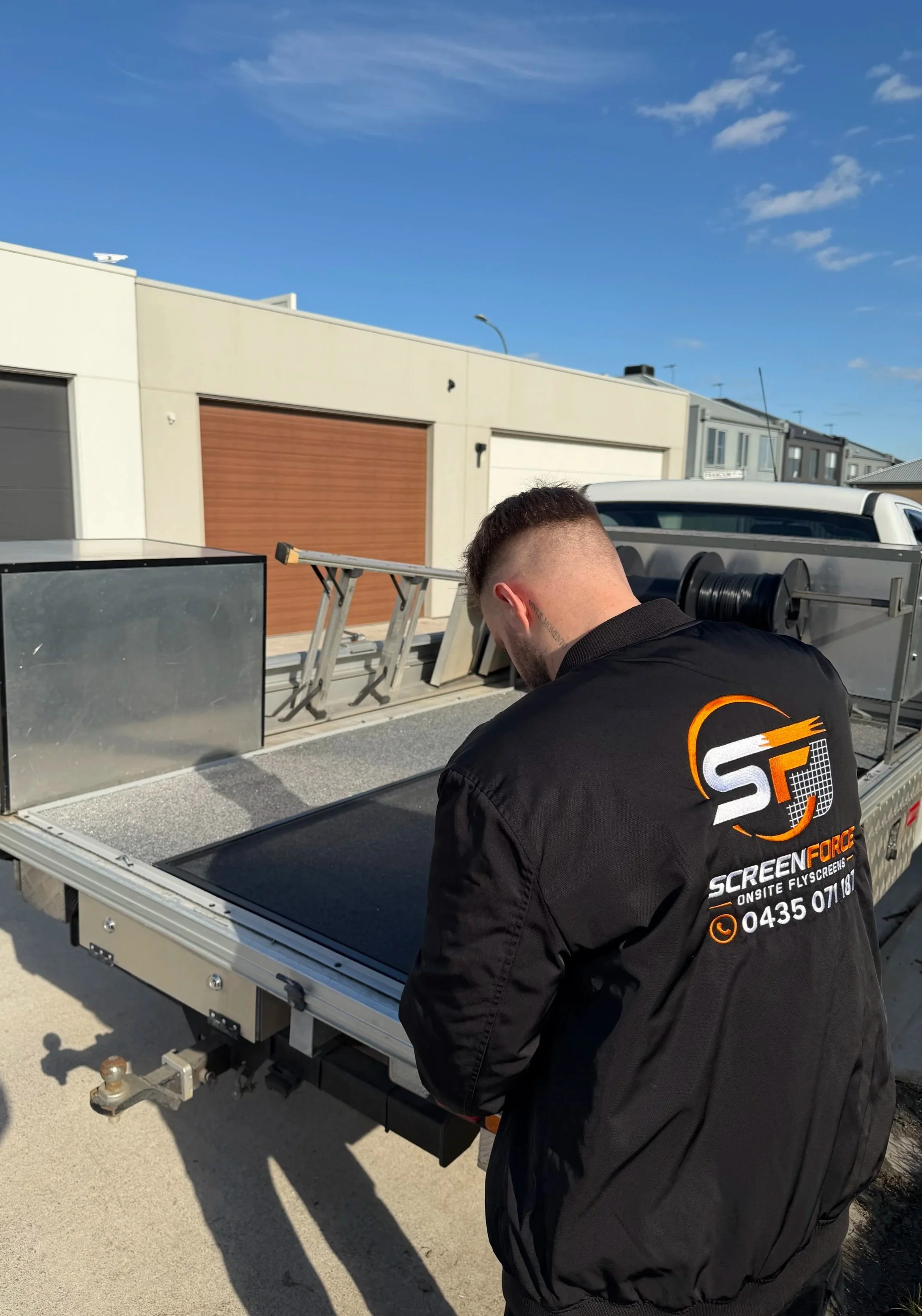 Person working on a mobile screen printing or signage setup on a trailer in a residential area with modern houses and blue sky in the background.