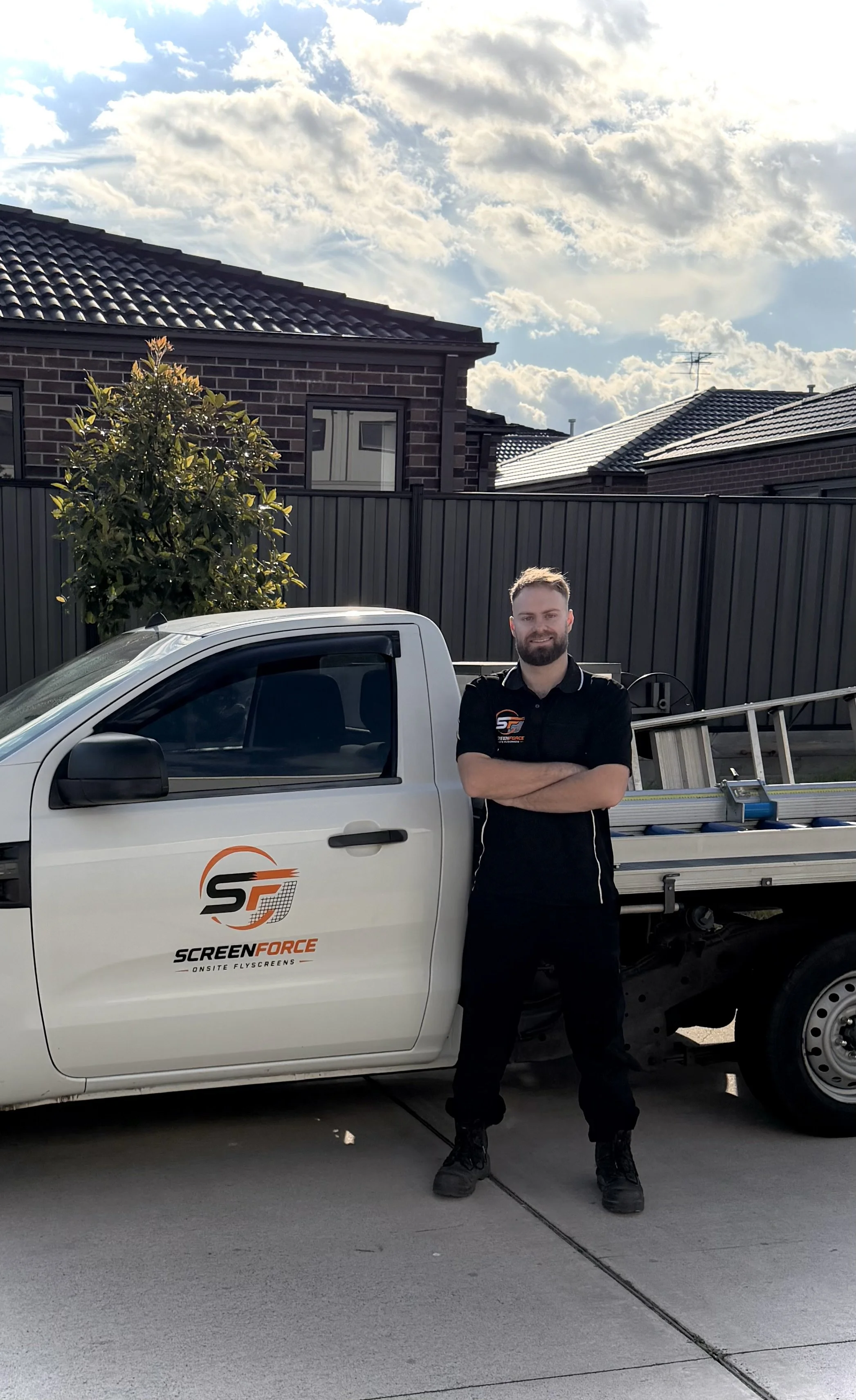A man with a beard wearing a black company shirt stands with arms crossed in front of a white service truck with the company logo 'ScreenForce'. The truck has a ladder on the back. The setting is a residential backyard with a black fence, trees, and houses under a partly cloudy sky.