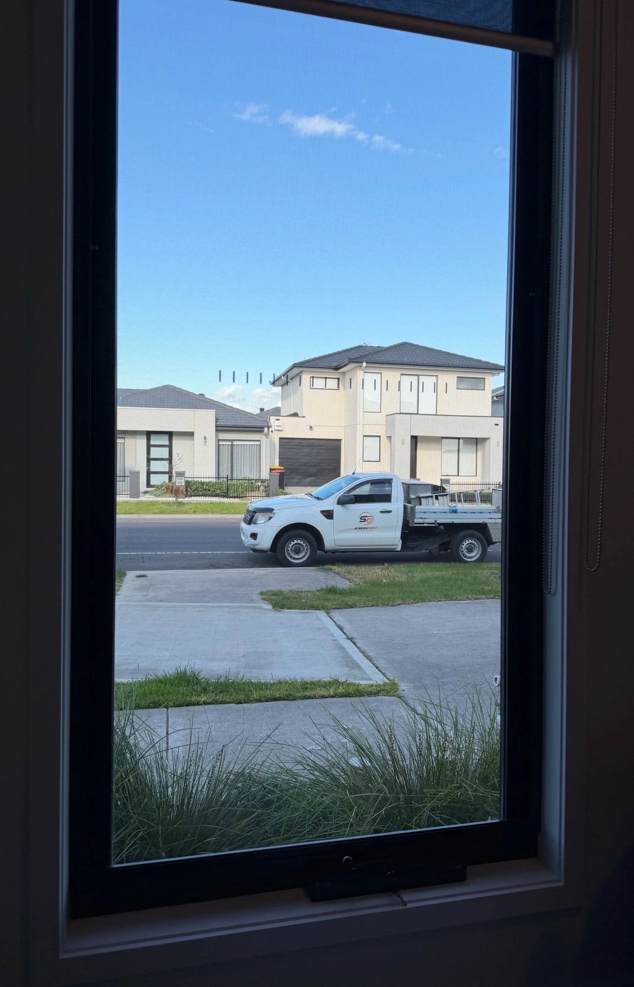 View through a window showing a white pickup truck parked on the street with a logo on its door, in front of modern residential houses with grey roofs and large windows, under a clear blue sky with a few clouds.