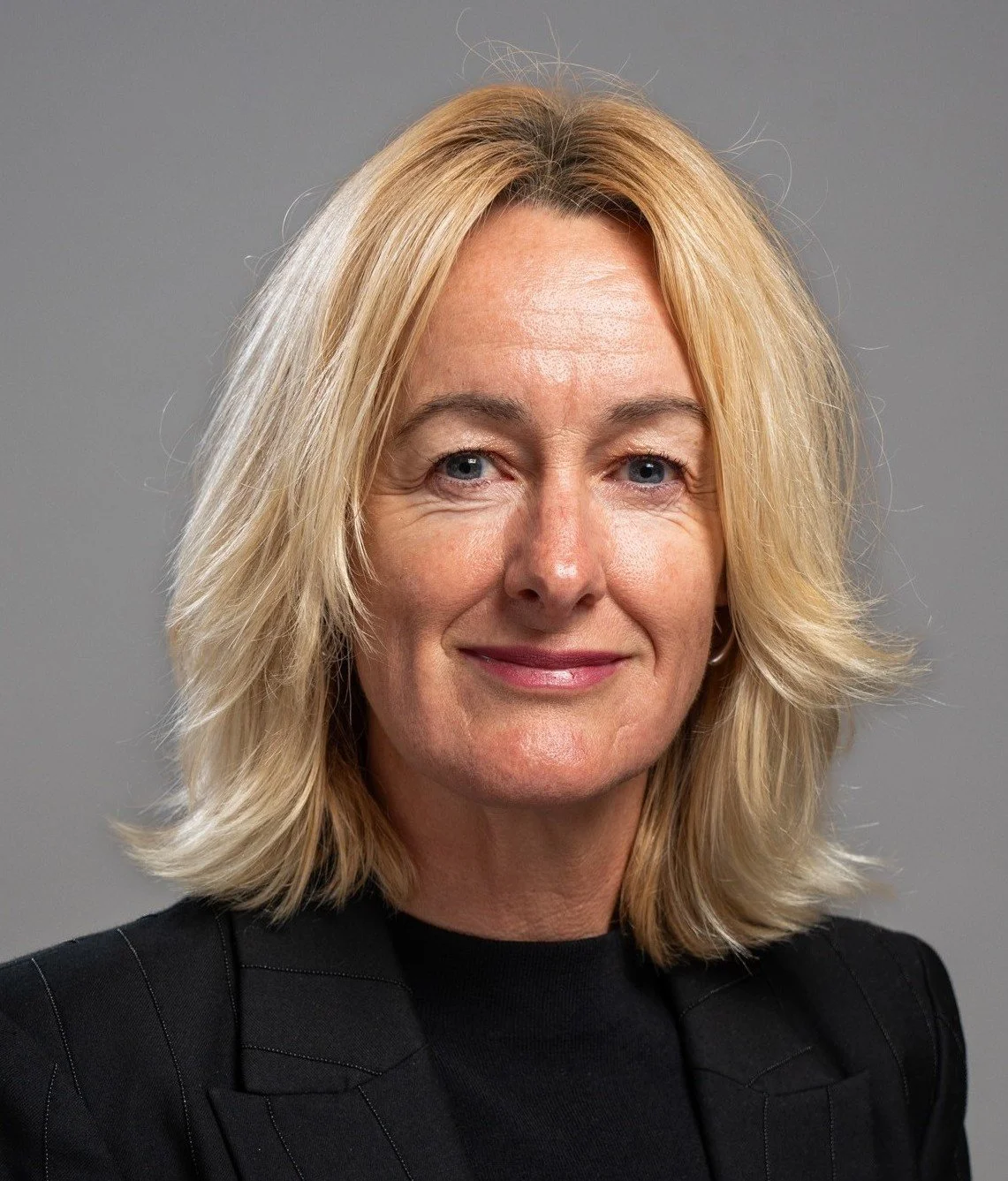 A middle-aged woman with shoulder-length blonde hair, blue eyes, and a slight smile, wearing a black blazer, against a gray background.