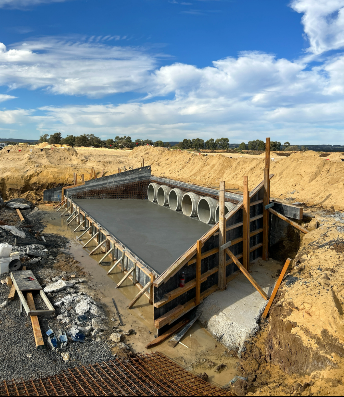 Construction site showing a concrete foundation with large pipes embedded, surrounded by dirt and wooden framing, under a partly cloudy sky.