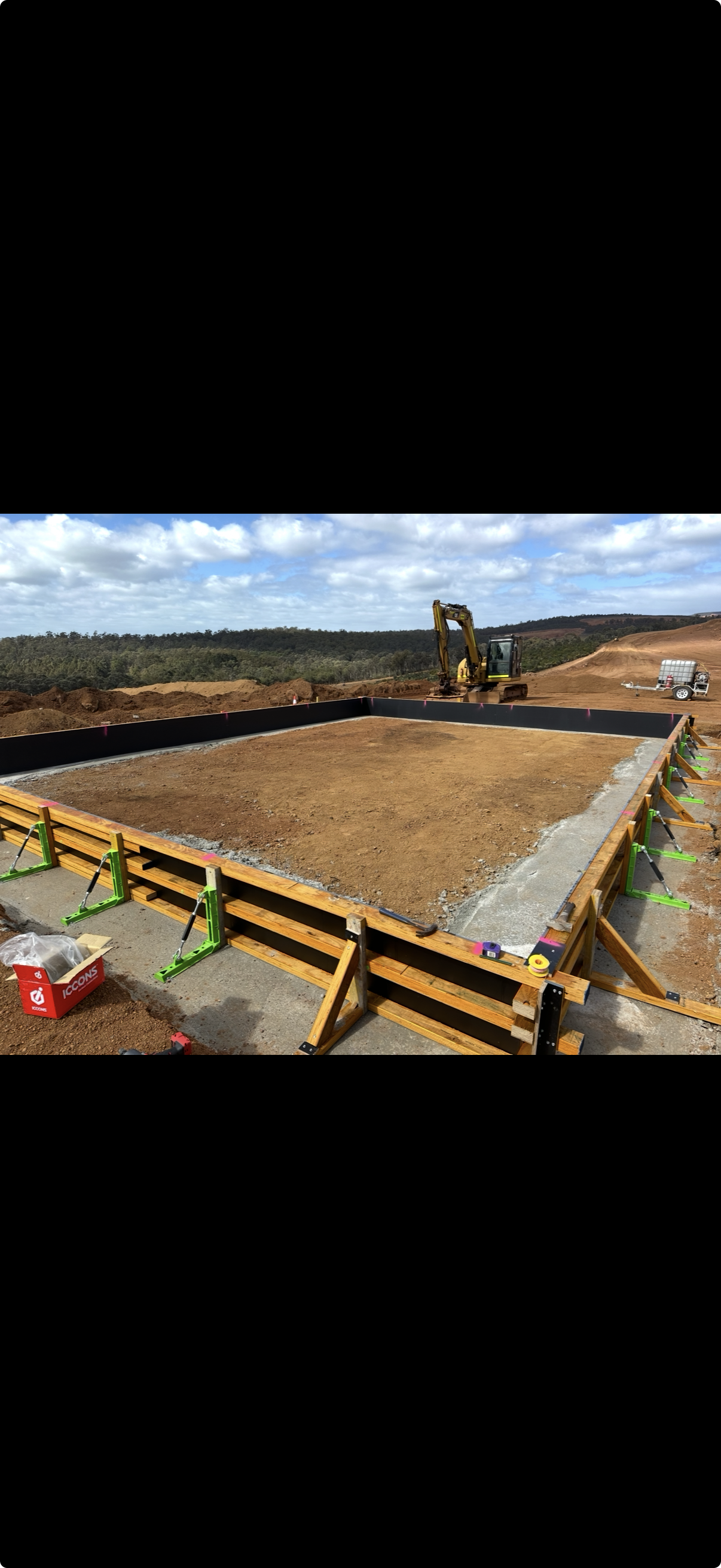Construction site with a large foundation being prepared, with a small excavator working inside the foundation perimeter, surrounded by safety barriers, with a clear sky and distant hills in the background.