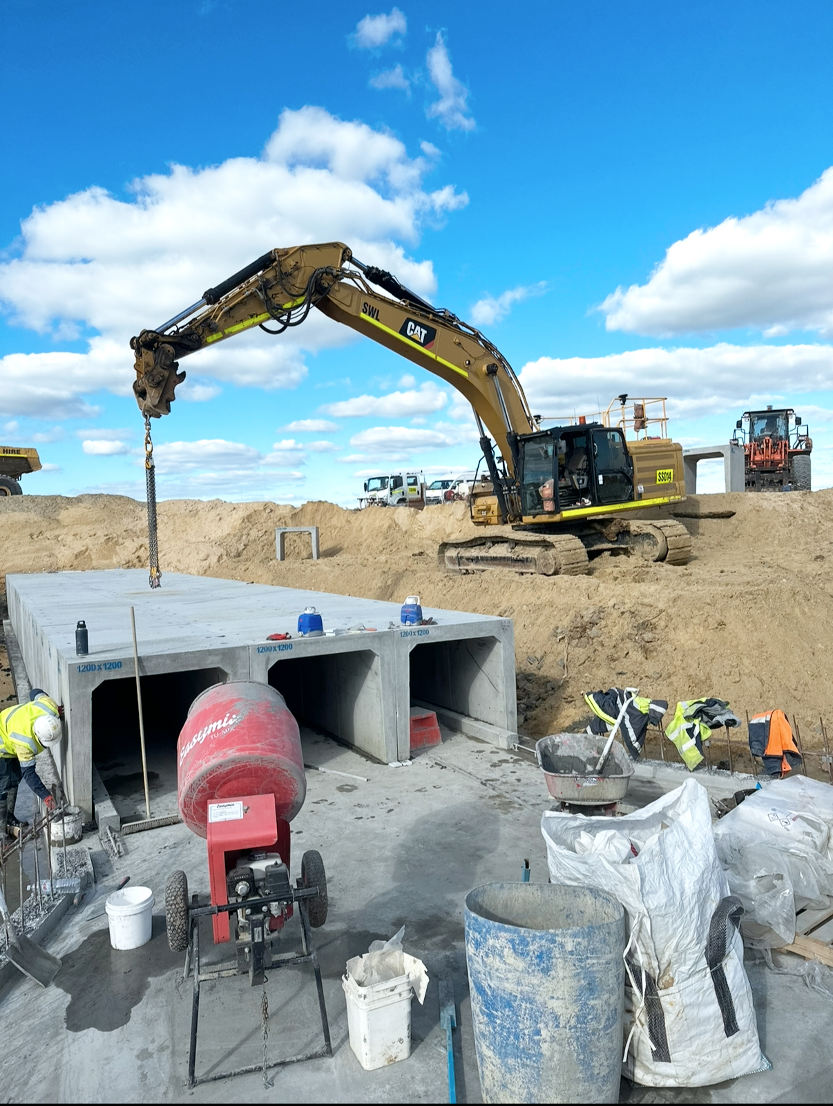 Construction site with excavator lifting a large concrete slab, workers in safety gear, various construction materials, and machinery against a bright blue sky with clouds.