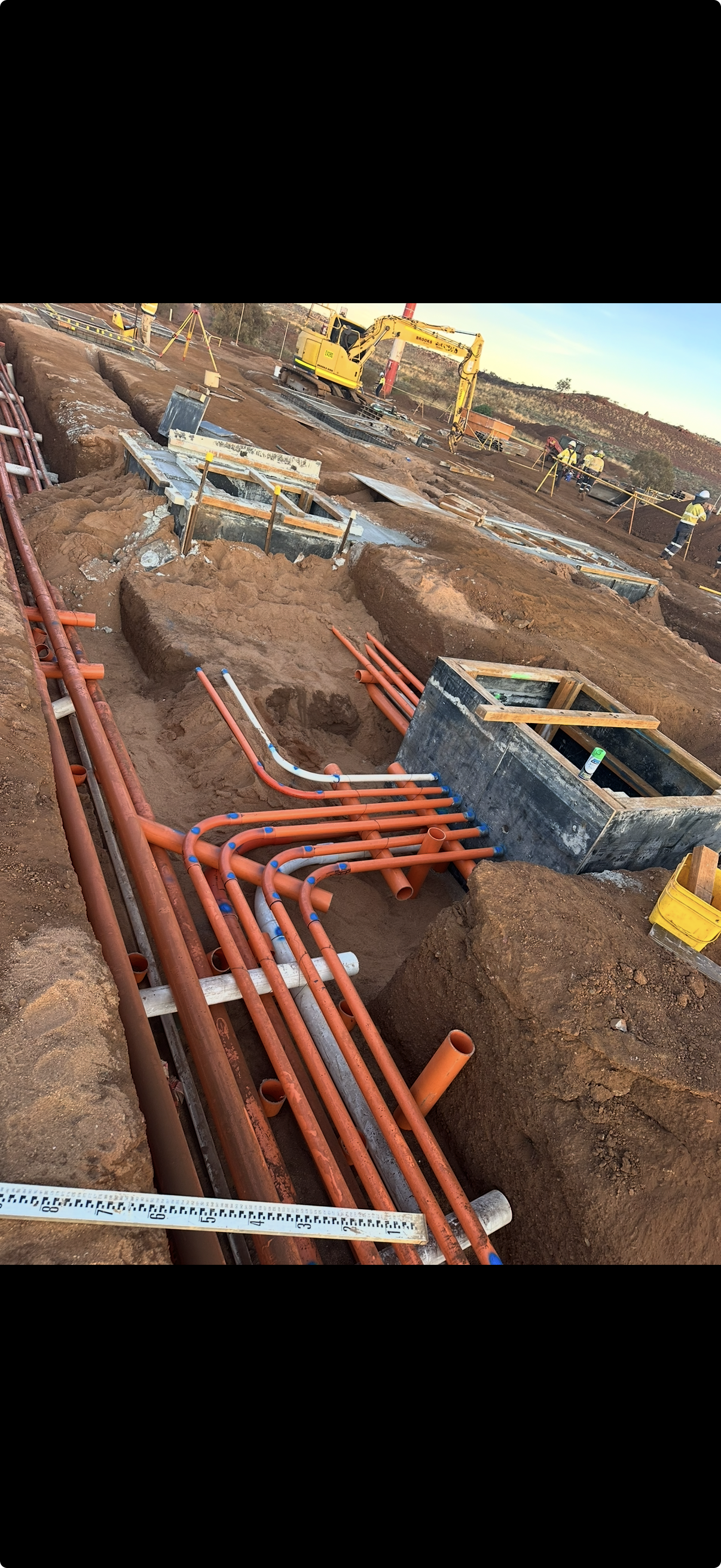 Construction site with exposed underground pipes and excavation trenches, with construction workers and heavy machinery in the background.