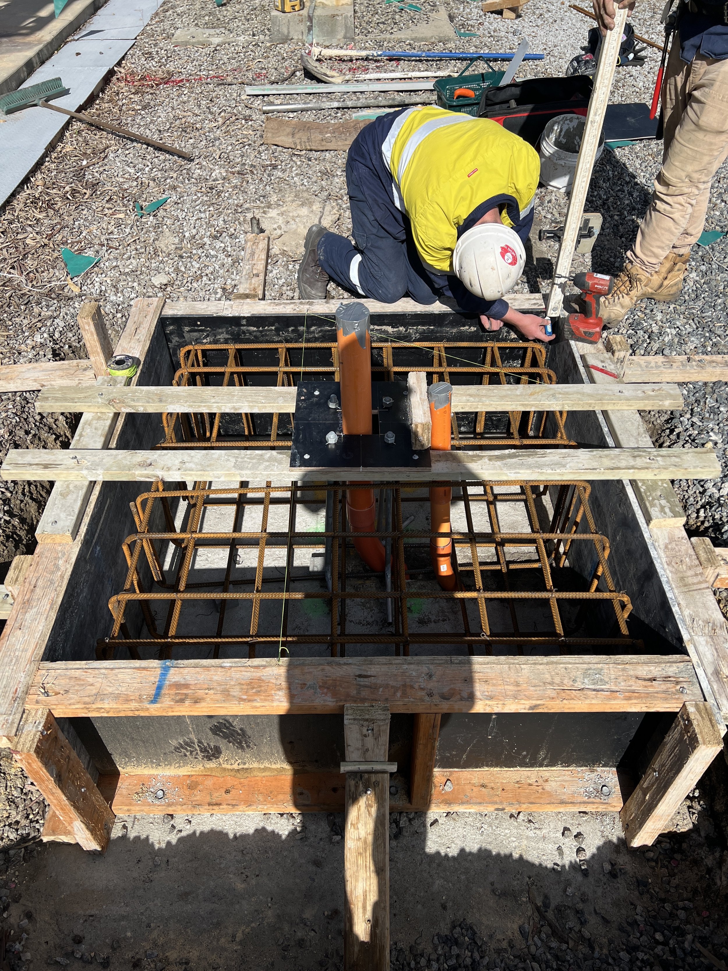 Construction site showing a worker kneeling and measuring inside a rectangular formwork reinforced with steel rebar. The worker is wearing a white helmet and a yellow safety vest. Surrounding the formwork are tools and construction materials.