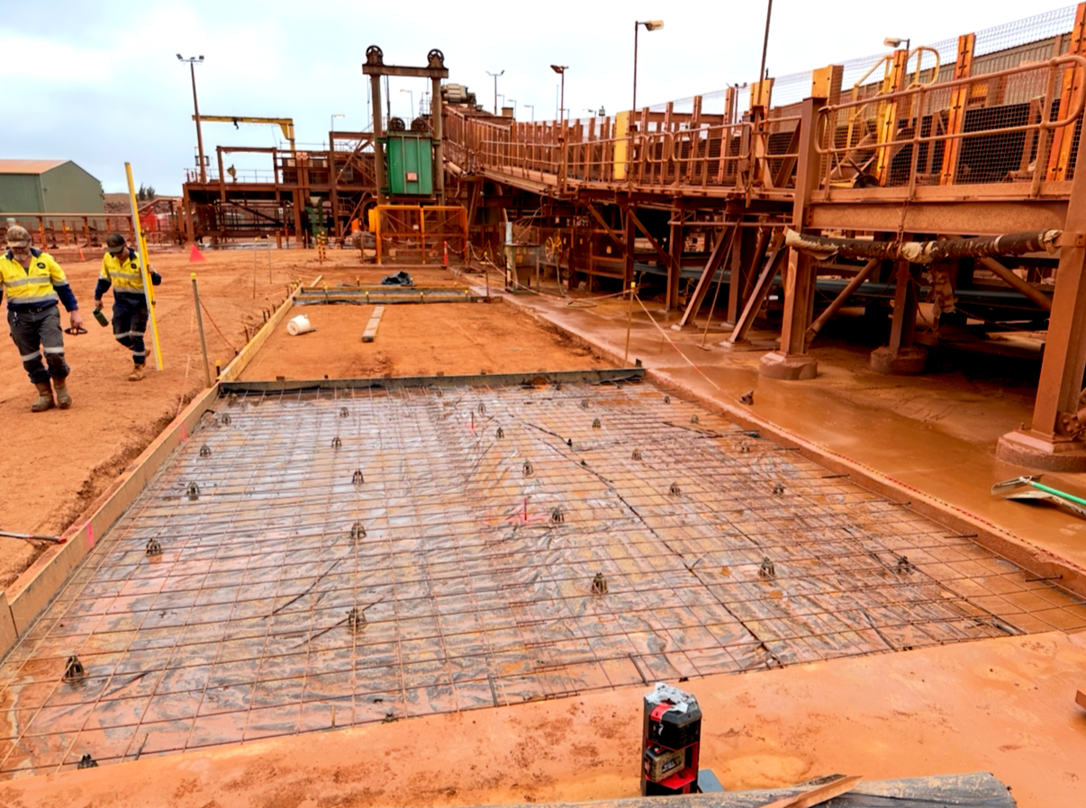 Construction workers walking on a construction site with steel reinforcement on the ground, machinery, and structures under construction.